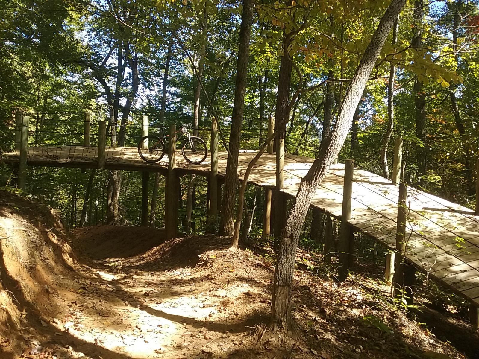 A mountain bike parked on a wooden bridge in a forested area, surrounded by trees with autumn foliage. The bridge is elevated above a dirt path that curves away from the viewer, emphasizing the natural setting. Sunlight filters through the leaves, creating a dappled light effect on the ground. Fire Mountain Trail System mountain bike trail.