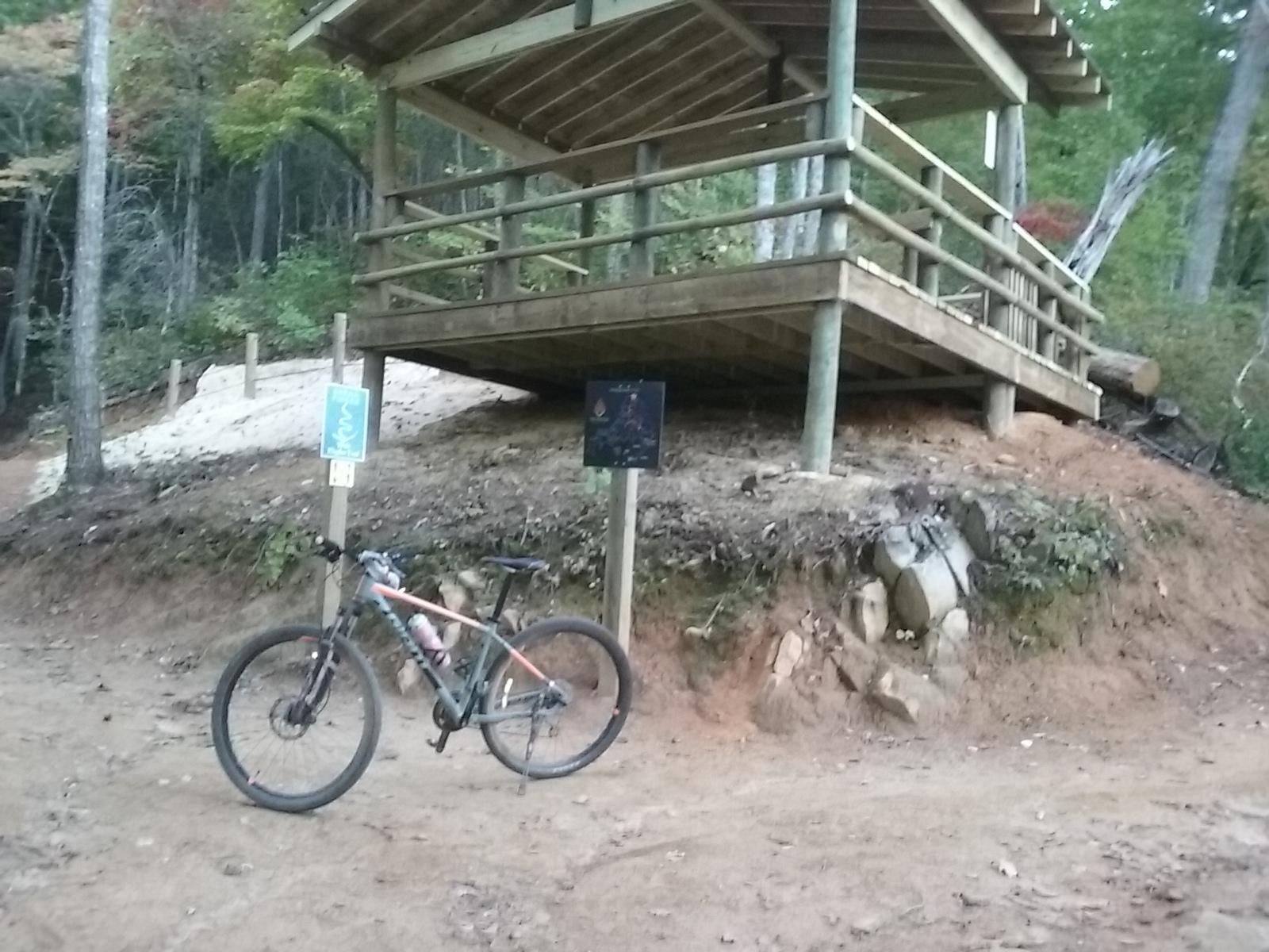 A mountain bike is parked on a dirt path next to a wooden shelter in a forested area. The shelter has a sloped roof and is supported by wooden posts. A sign is visible nearby, along with a designated bike trail marker. Surrounding greenery indicates a natural, outdoor setting. Fire Mountain Trail System mountain bike trail.
