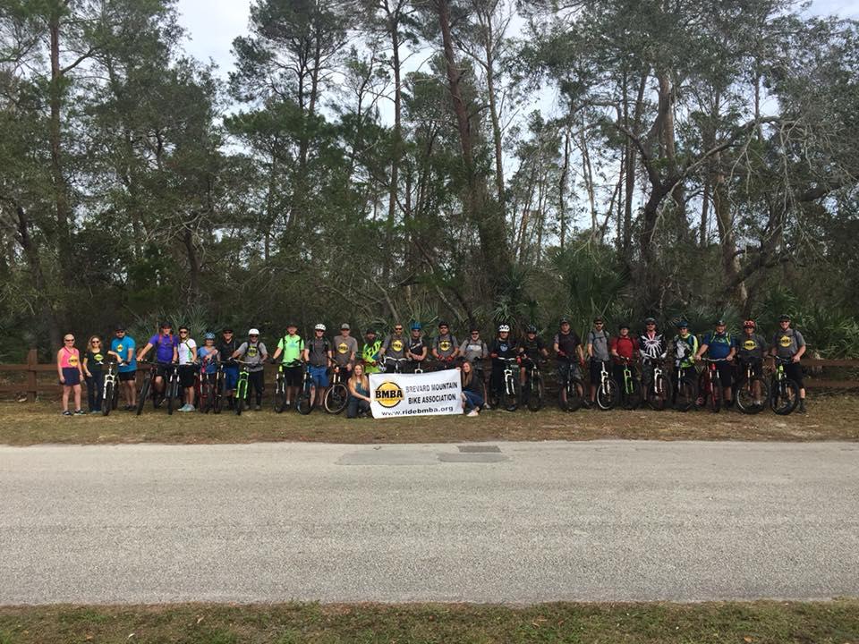 A group of mountain biking enthusiasts stands together with their bikes in a wooded area. They are posing in front of a wooden fence, holding a banner for the Brevard Mountain Bike Association. The group comprises various age ranges and genders, wearing cycling attire and helmets, surrounded by trees and greenery.