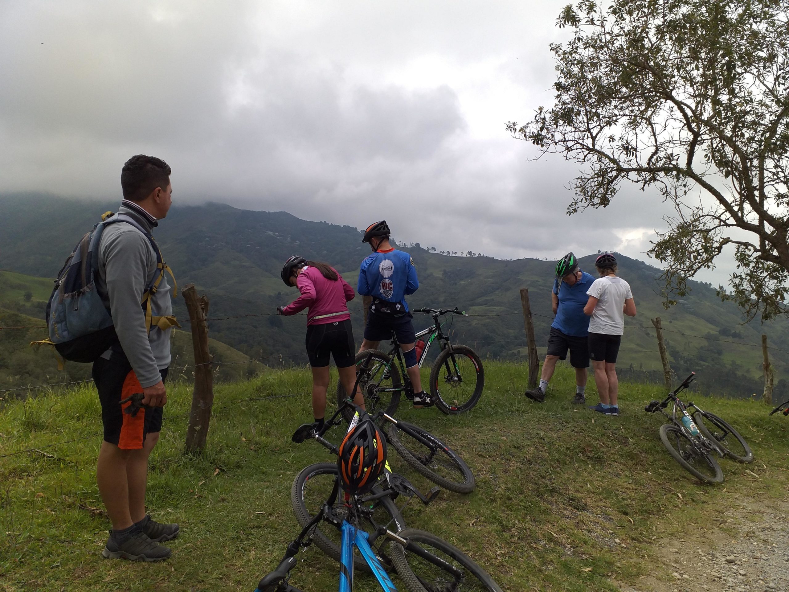A group of five cyclists stop to rest on a hillside overlooking a scenic green landscape. Two cyclists are adjusting their gear, while another cyclist stands nearby, looking out at the view. Their bicycles are parked on the grass, and the sky is partly cloudy, creating a serene outdoor atmosphere. Alegria mountain bike trail.