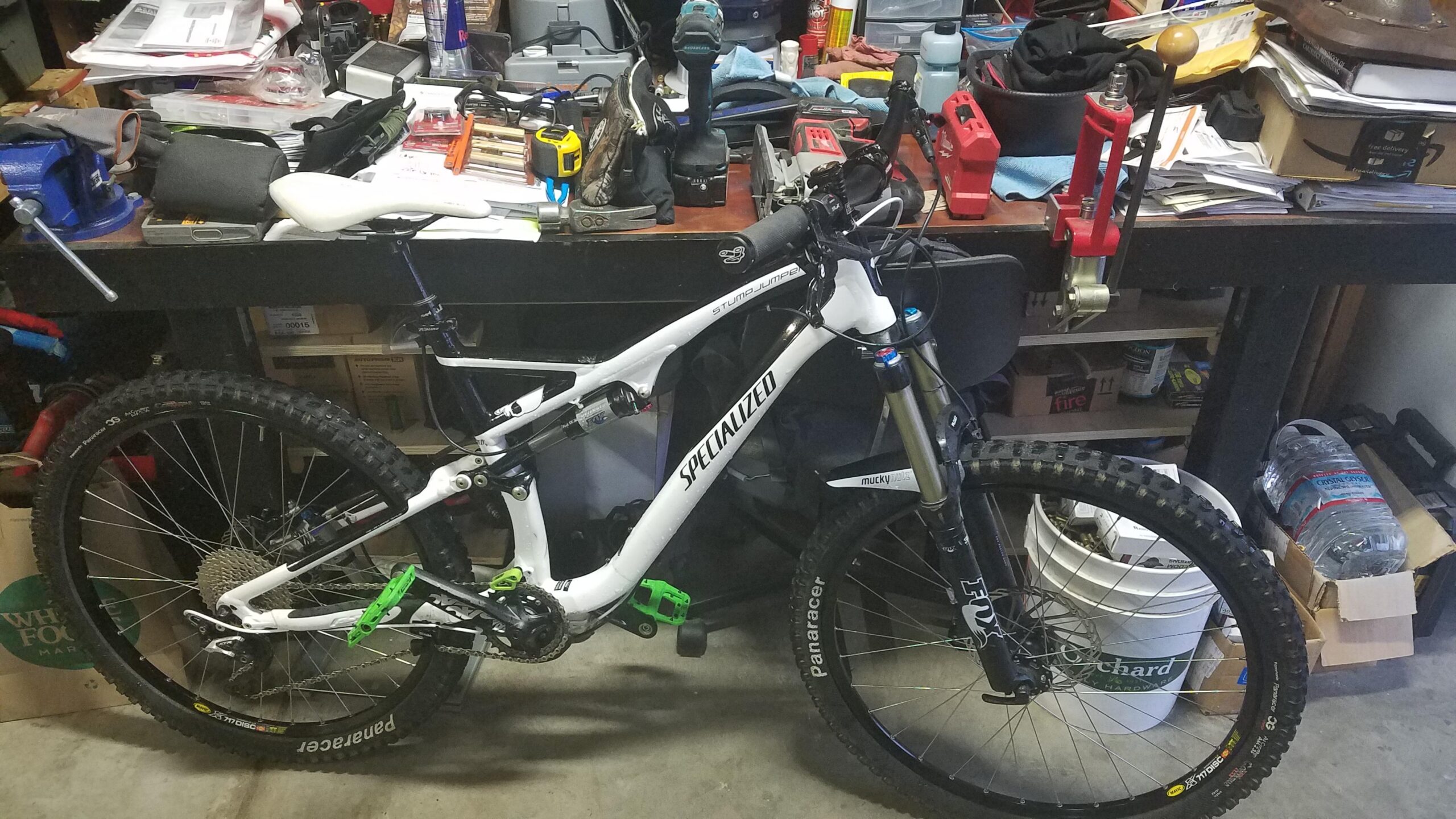 Specialized Stumpjumper FSR Elite: Alt text: A white and black mountain bike, specifically a Specialized Stumpjumper, is leaning against a cluttered workbench filled with tools, paperwork, and various items. The bike features thick tires and green pedal clips. In the background, there are bins and containers, adding to the workshop setting.