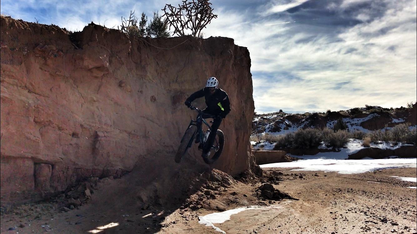 A mountain biker performing a jump near a rocky cliff in a desert landscape, with scattered snow and sparse vegetation in the background. The sky is partly cloudy, creating a dramatic atmosphere. Mariposa Fat Bike Trails mountain bike trail.