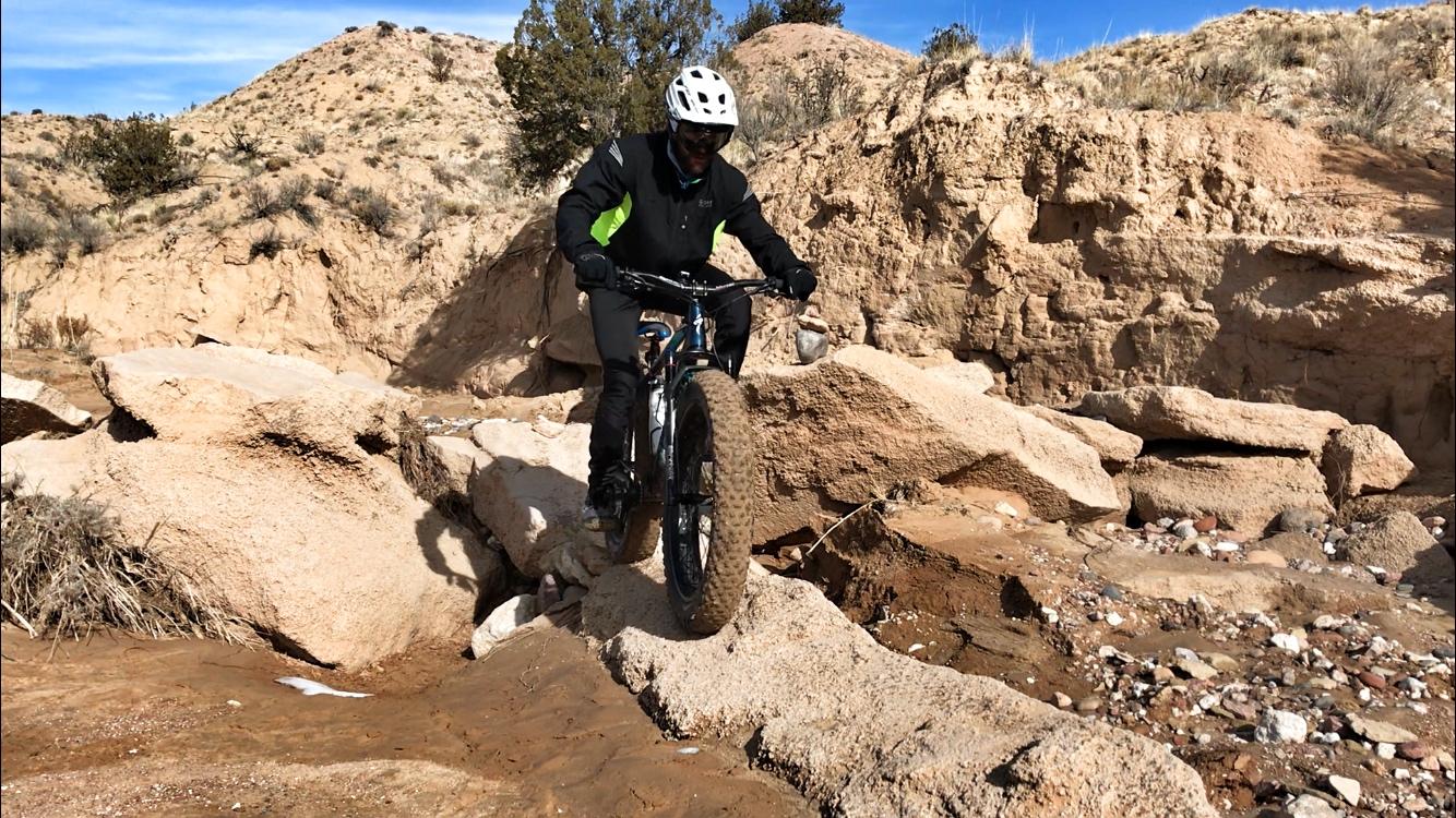 A person riding a mountain bike over rocky terrain in a desert landscape, wearing a helmet and dark clothing, with a backdrop of sandy hills and sparse vegetation. Mariposa Fat Bike Trails mountain bike trail.