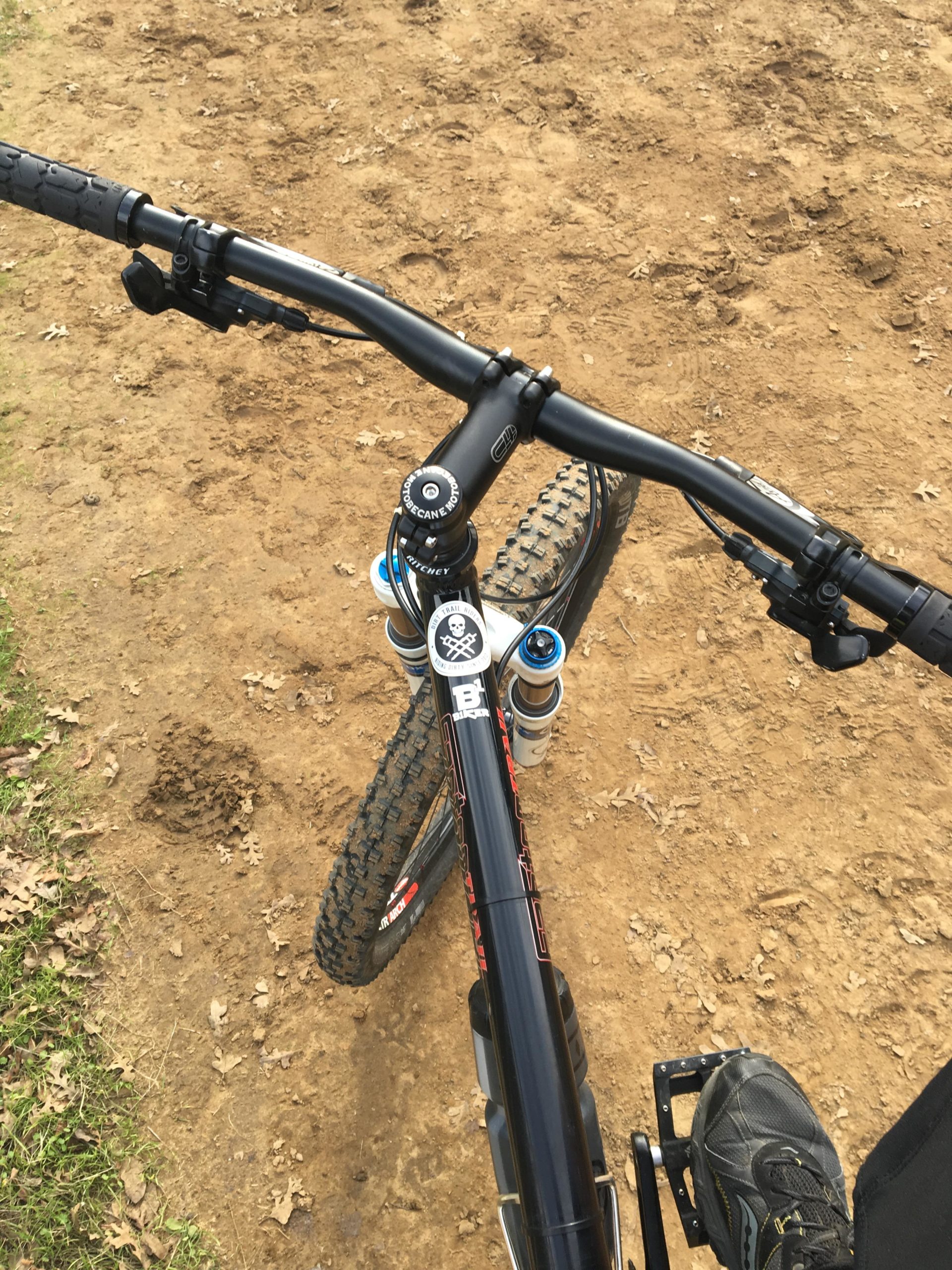 A top-down view of a mountain bike handlebars, showcasing the grip, brake lever, and suspension controls. The bike's front wheel is partially visible on the left, positioned on a dirt path with fallen leaves scattered around. There is a foot resting on the pedal, indicating readiness to ride. Round Valley to Morgan Territory mountain bike trail.