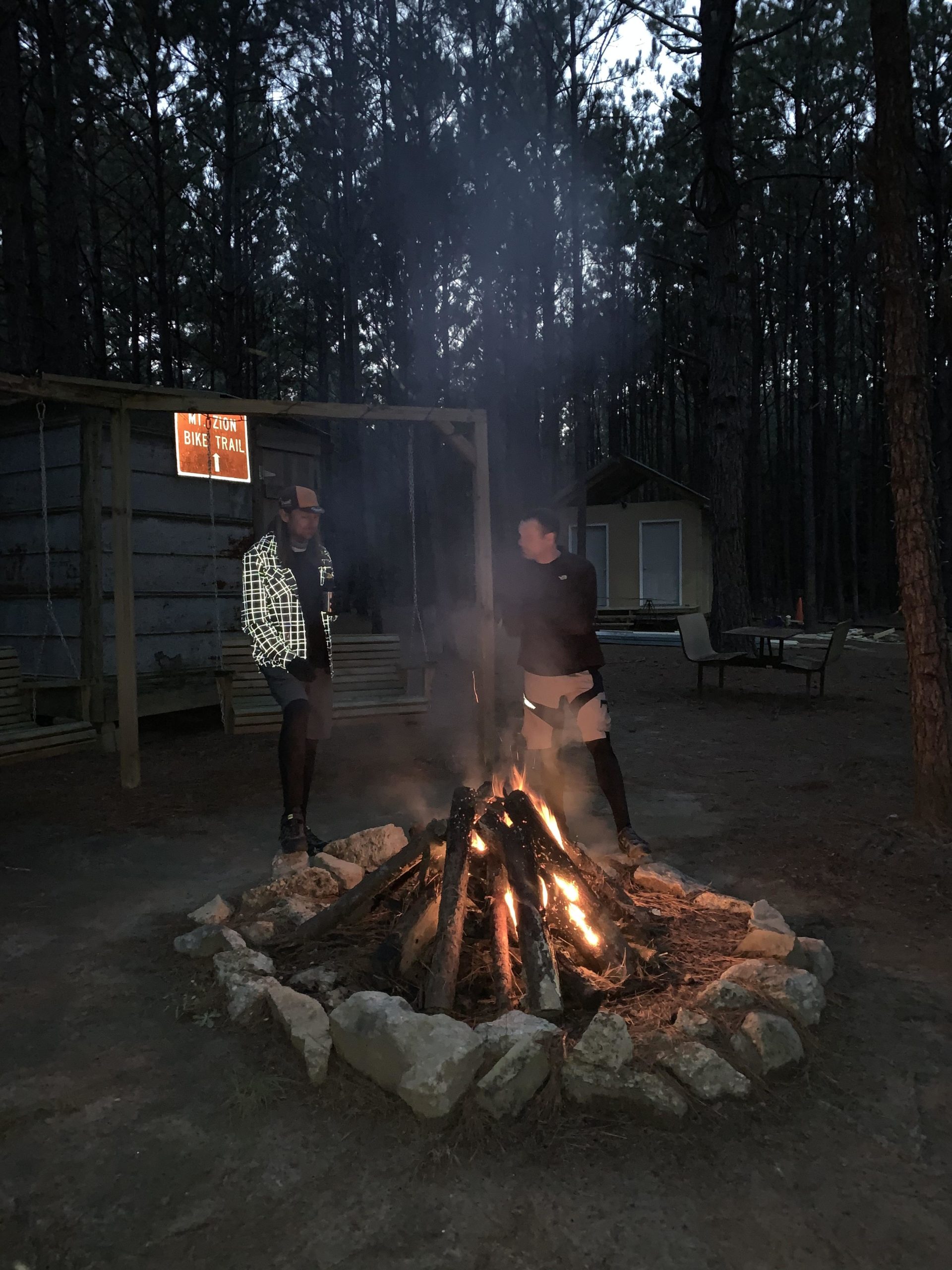 Two people stand around a campfire in a wooded area during dusk. The campfire is surrounded by a circle of stones, with flames and smoke rising into the air. In the background, a sign indicates the "Zion Bike Trail," and there are trees surrounding the campsite. A few outdoor furniture pieces are visible to the side. Mt. Zion Bike Trails mountain bike trail.