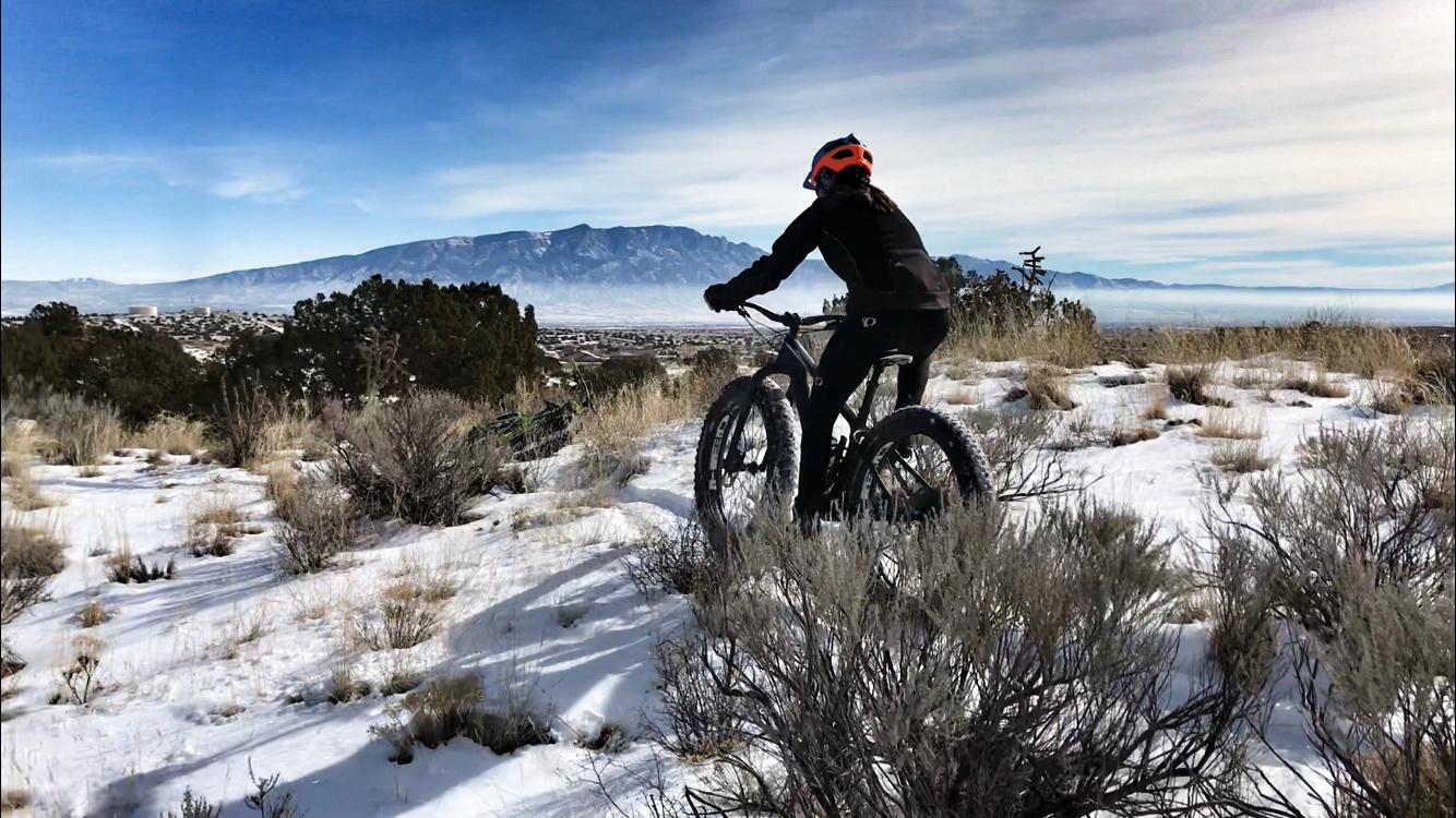 A person riding a fat tire bike on a snowy trail, surrounded by sparse vegetation and distant mountains under a clear blue sky. The scene captures a sense of adventure and exploration in a winter landscape. Mariposa Fat Bike Trails mountain bike trail.