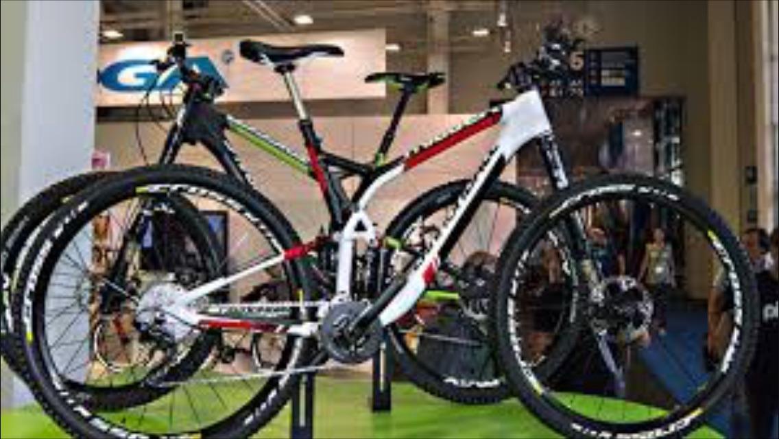 Cannondale Trigger Carbon 2: Two mountain bikes displayed at a trade show booth, one with a white frame featuring red and black accents, and the other with a black frame highlighted in green. Both bikes are positioned on a bright green platform, showcasing their wheels and design against a blurred background of event attendees and promotional signage.