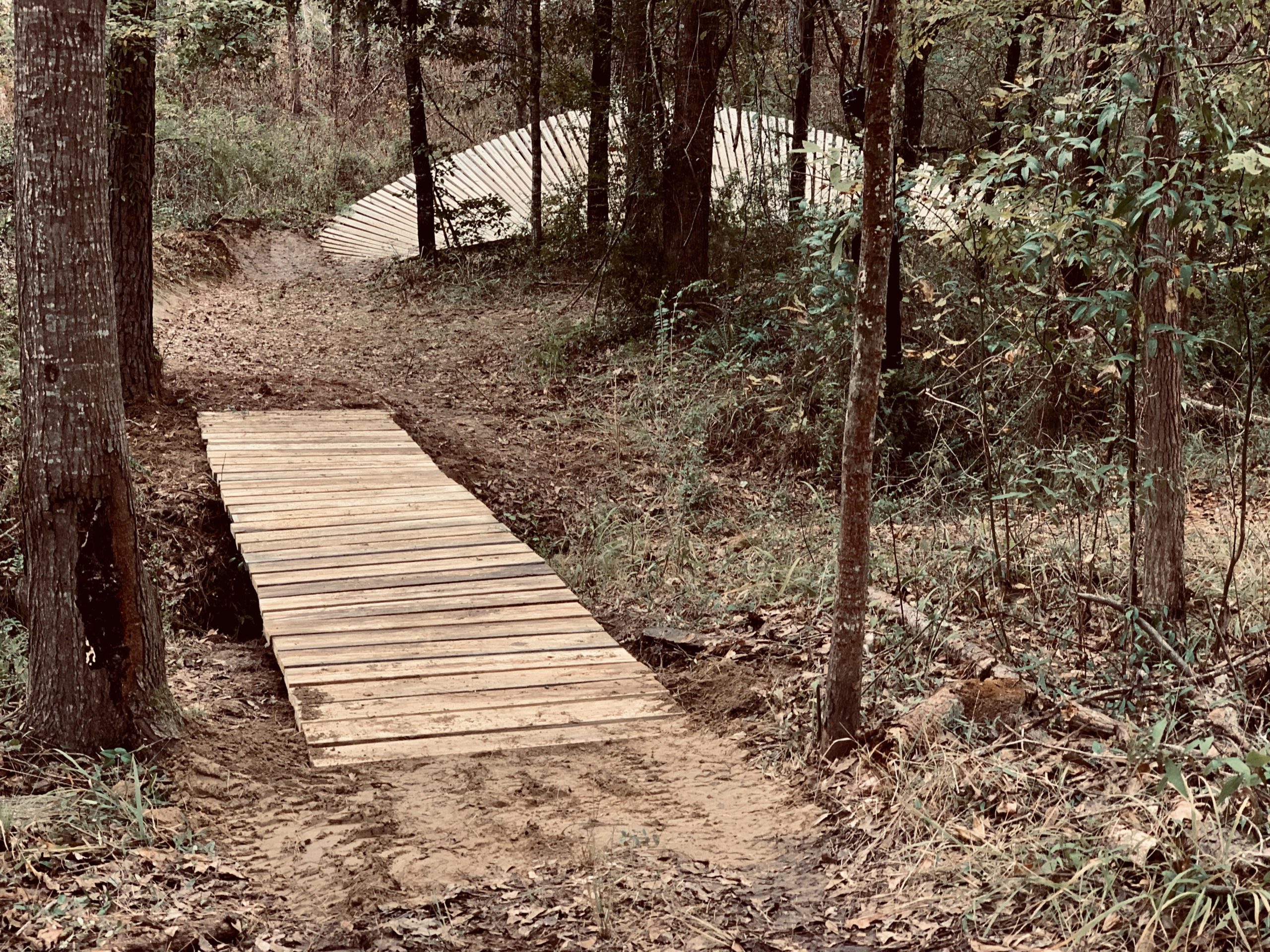 A wooden bridge spans a dirt path in a forested area, surrounded by trees and underbrush. In the background, a slanted wooden structure can be seen, blending into the natural setting. The ground is covered with fallen leaves and the scene is tranquil, suggesting a rural or hiking trail environment. Mt. Zion Bike Trails mountain bike trail.