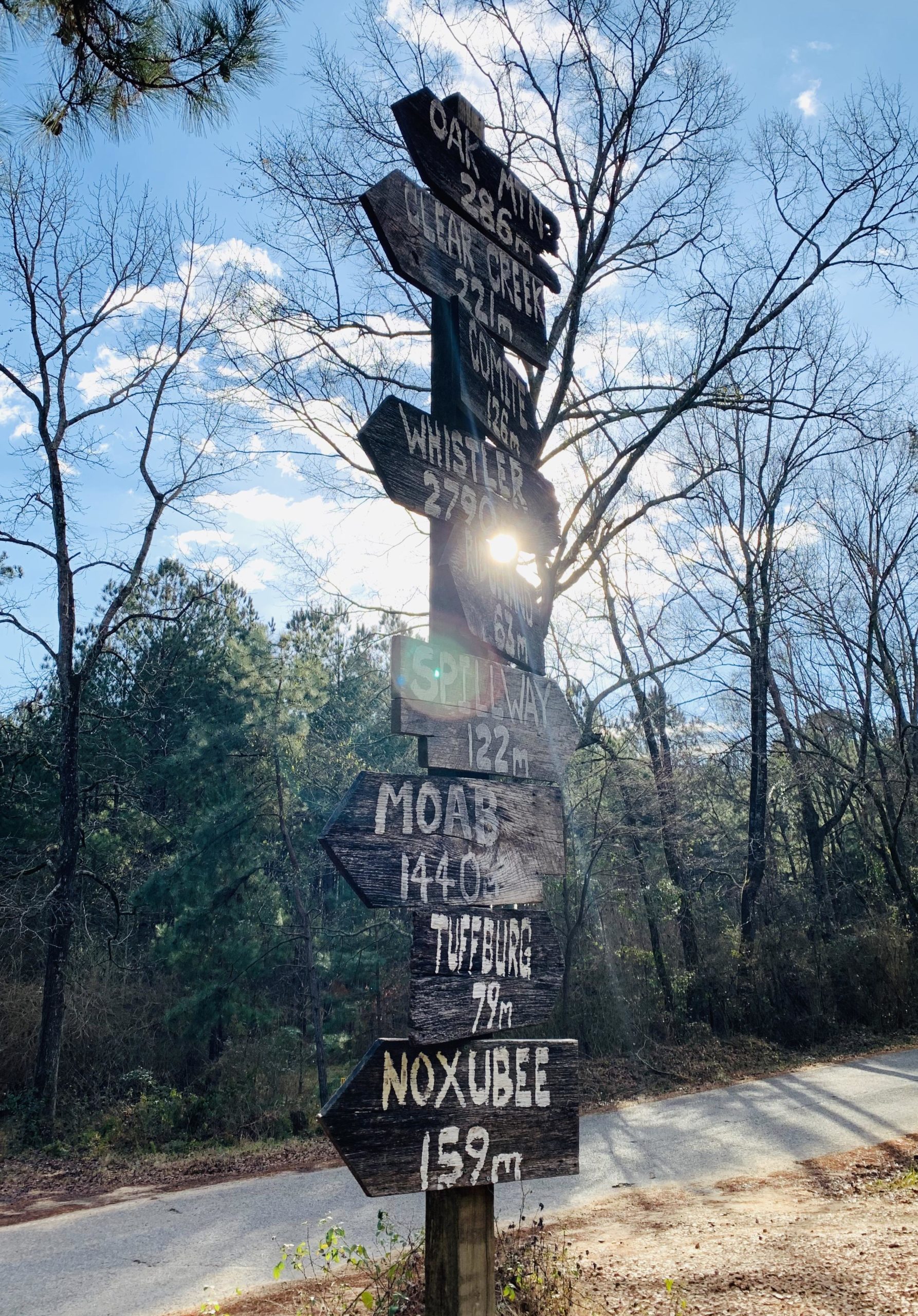 A weathered wooden signpost with multiple directional arrows pointing towards various locations and distances, set against a blue sky with scattered clouds and trees in the background. The sign indicates distances to destinations such as Oak Mountain, Clear Creek, and Moab, among others. Sunlight shines through the sign, creating a glowing effect. Mt. Zion Bike Trails mountain bike trail.
