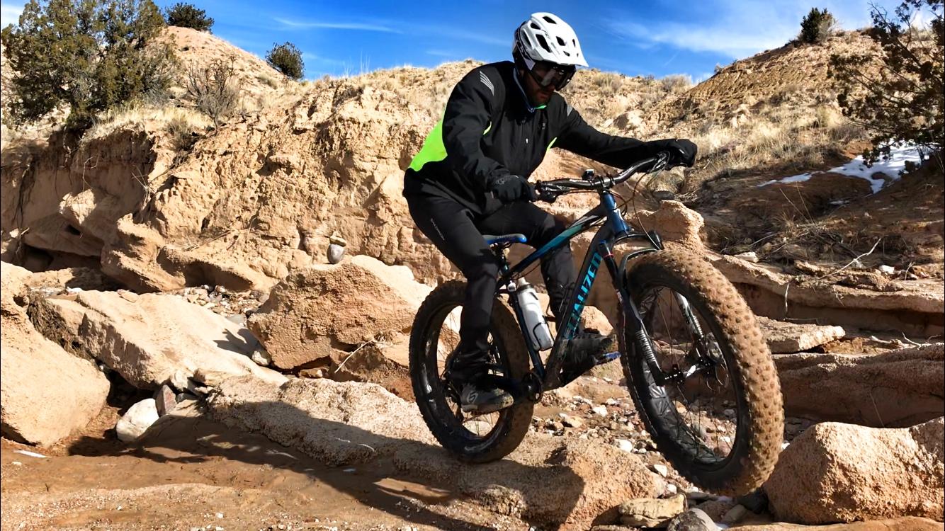 A cyclist wearing a black and neon green jacket and helmet rides a fat bike over rocky terrain, with hills and sparse vegetation in the background under a clear blue sky. Mariposa Fat Bike Trails mountain bike trail.