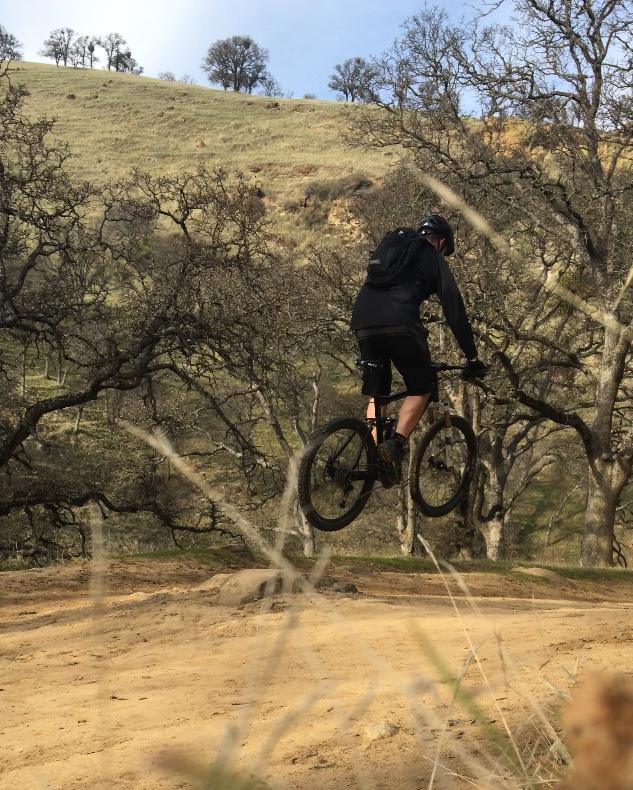 A mountain biker in a black jersey and shorts performs a jump off a dirt ramp surrounded by trees and a hilly landscape. The cyclist is airborne, with a focus on maintaining balance and control. The background features grassy hills and a clear blue sky. Round Valley to Morgan Territory mountain bike trail.
