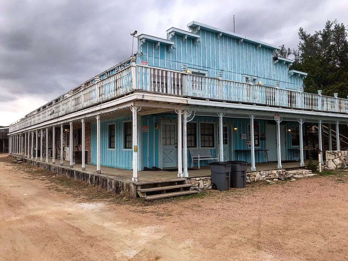 A light blue, two-story building with a wraparound porch, located in a rustic environment. The structure features white railings, large windows, and multiple doors. The surrounding area is sandy, with a few trees visible in the background under a cloudy sky. Saloon mountain bike trail.