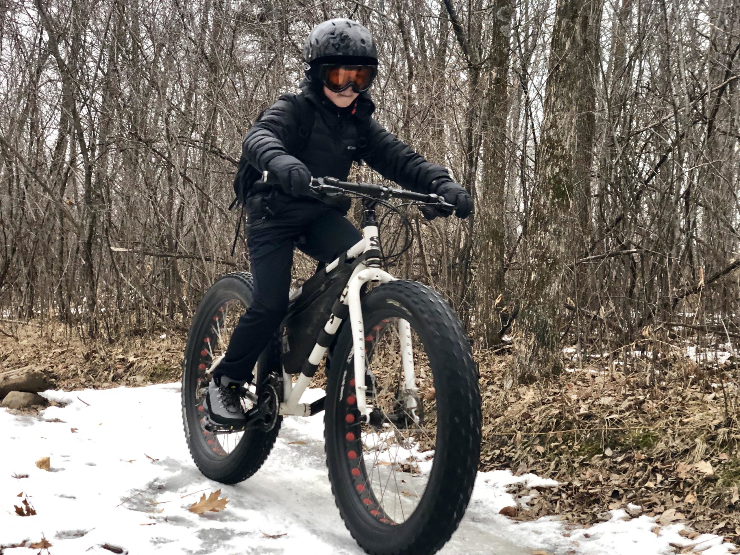 A person riding a fat bike on a snow-covered trail surrounded by trees in a winter landscape. They are wearing a black helmet and goggles, along with a black jacket and gloves. Elm Creek Park mountain bike trail.