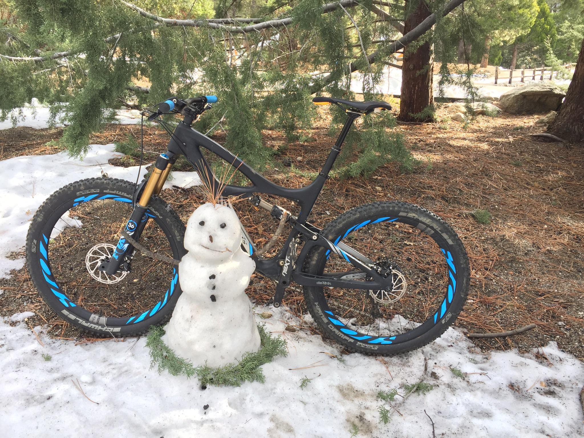 Ibis Mojo HD4: A mountain bike leaning against a snowman in a forested area, with patches of snow on the ground and pine needles scattered around. The snowman has small rocks for eyes and buttons, and is adorned with a sprig of pine on top.