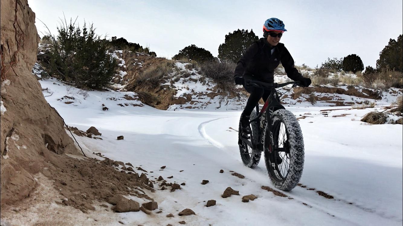 A person riding a fat bike on a snowy trail, surrounded by a hilly landscape and scattered vegetation. The cyclist is wearing a helmet and sunglasses, pedaling through the snow with tire tracks visible behind them. The scene conveys a sense of adventure in a winter setting. Mariposa Fat Bike Trails mountain bike trail.