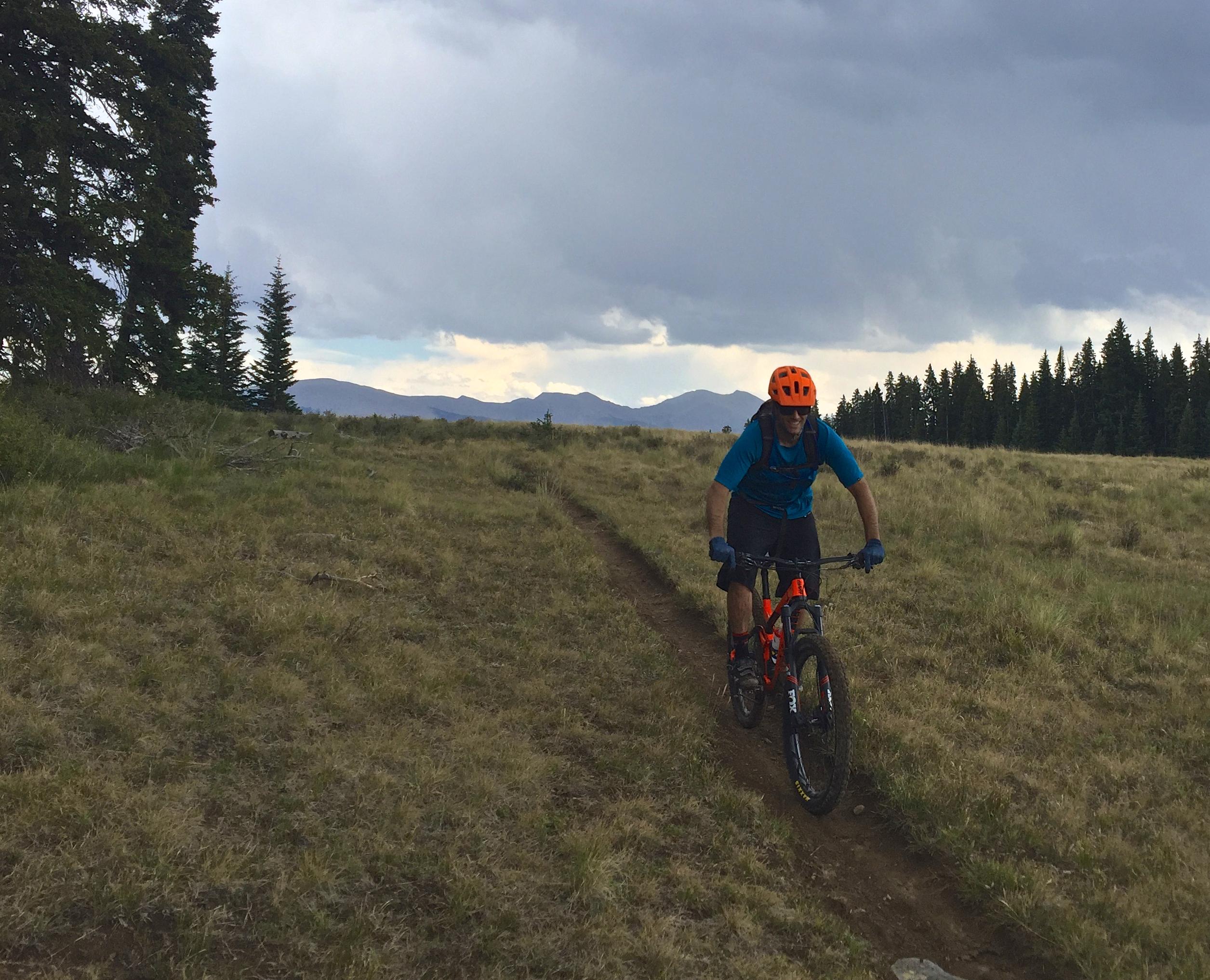 A mountain biker wearing an orange helmet and blue shirt navigates a dirt trail through a grassy landscape, with trees and distant mountains in the background under a cloudy sky. Doctor Park mountain bike trail.