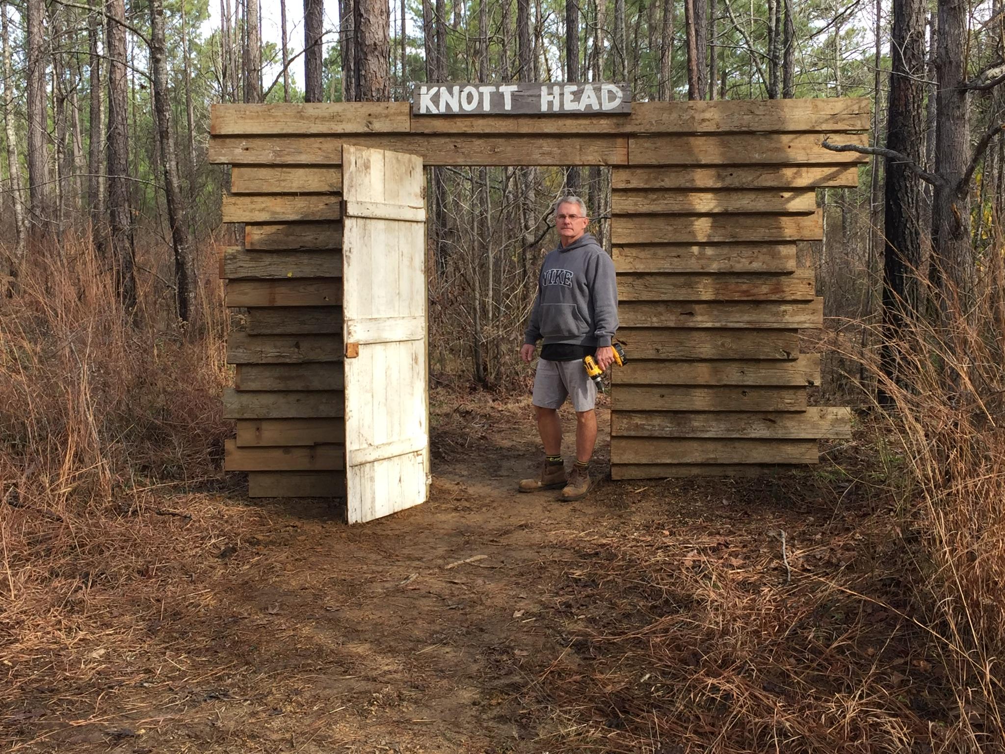 Alt text: A man stands next to a wooden structure labeled "KNOTT HEAD" in a forested area. The structure has an open door and is made from stacked wooden planks, with trees and tall grass surrounding it. The man is wearing a gray sweatshirt, shorts, and work boots, holding a power tool. Mt. Zion Bike Trails mountain bike trail.