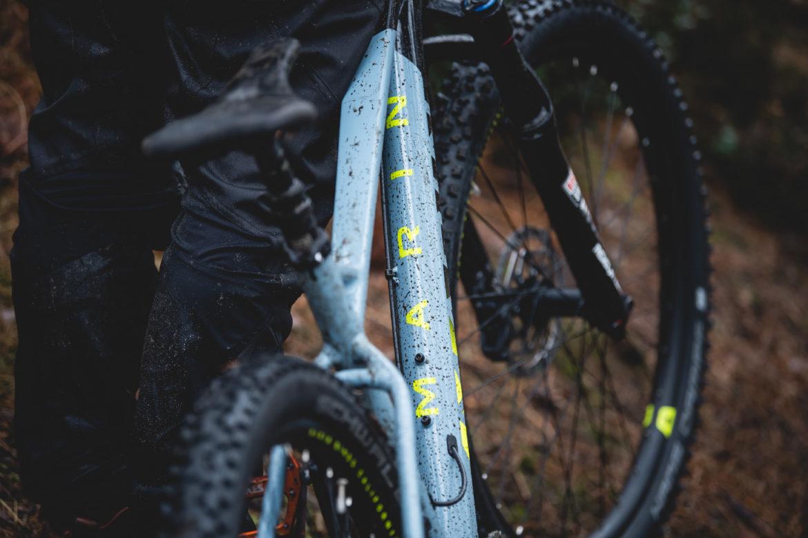 Marin San Quentin 2: A close-up of a mountain bike with a blue frame, partially covered in mud, showcasing its yellow branding. The bike's seat and rear wheel are visible, while a person in dark pants stands next to it, hinting at outdoor riding conditions.