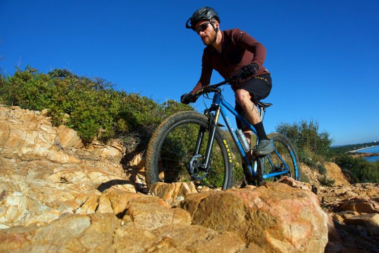 A mountain biker navigates a rocky terrain under a clear blue sky, showcasing the thrill of outdoor adventure. The cyclist is wearing a helmet and sunglasses, dressed in a long-sleeved top and shorts. Surrounding foliage adds a vibrant green contrast to the rocky landscape.
