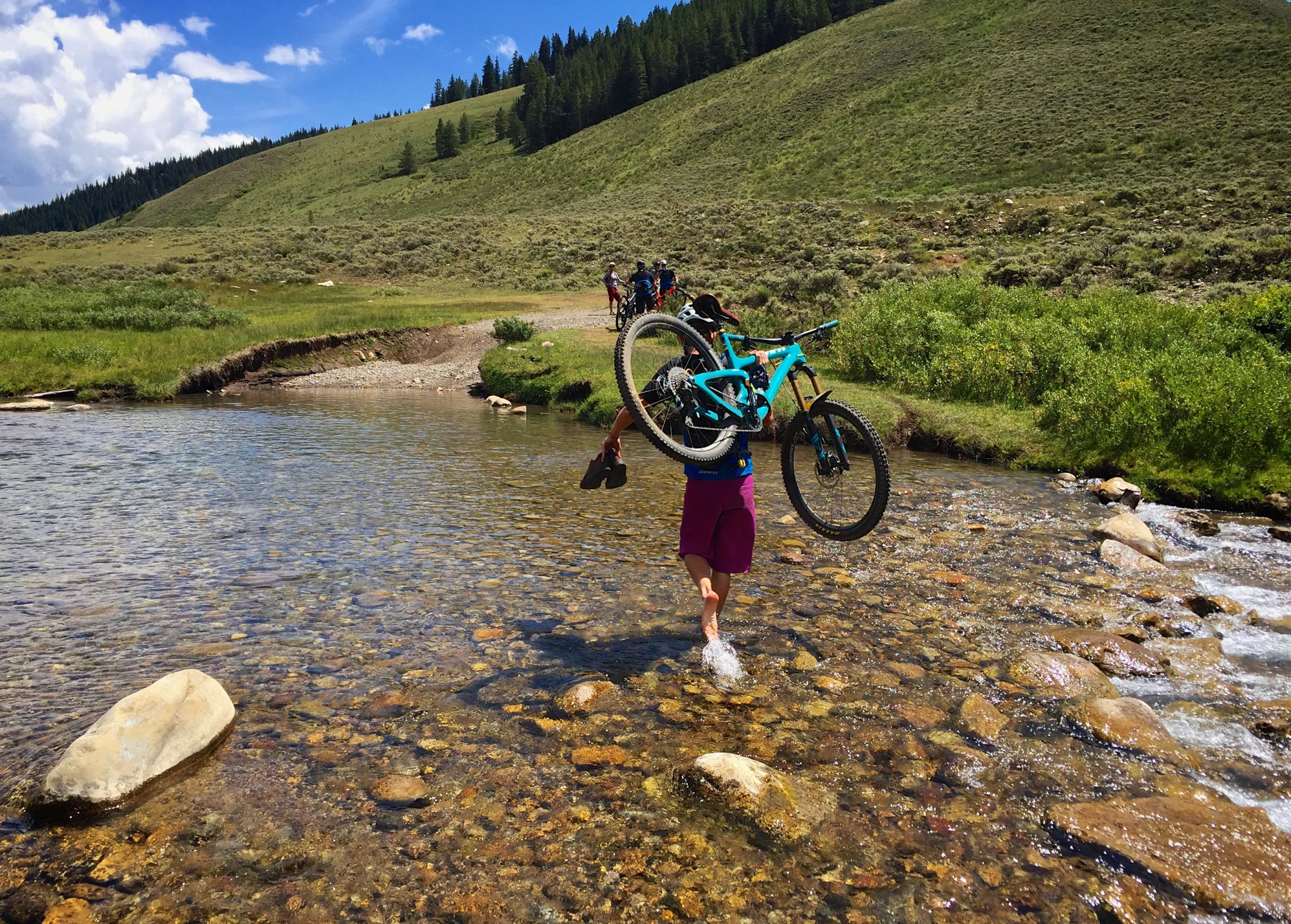 A person wades through a shallow stream, carrying a mountain bike over their shoulder and holding shoes in one hand. Lush green hills and a blue sky with scattered clouds are visible in the background, along with a few other people and bikes in the distance. The scene showcases an adventurous outdoor activity in a beautiful natural setting. Doctor Park mountain bike trail.