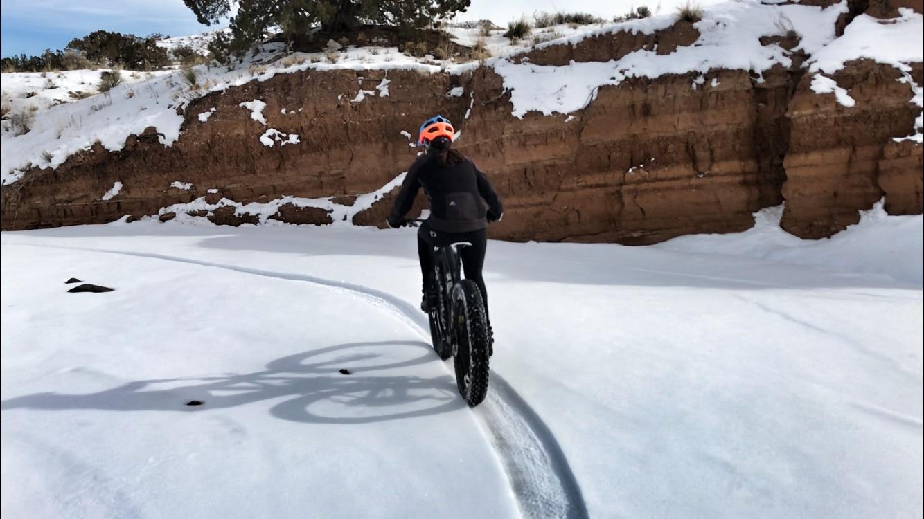 A person riding a fat bike through a snowy landscape, with a rocky cliff in the background. The snow is undisturbed except for the tire tracks left by the bike. The scene is set under a clear blue sky, indicating a bright winter day. Mariposa Fat Bike Trails mountain bike trail.