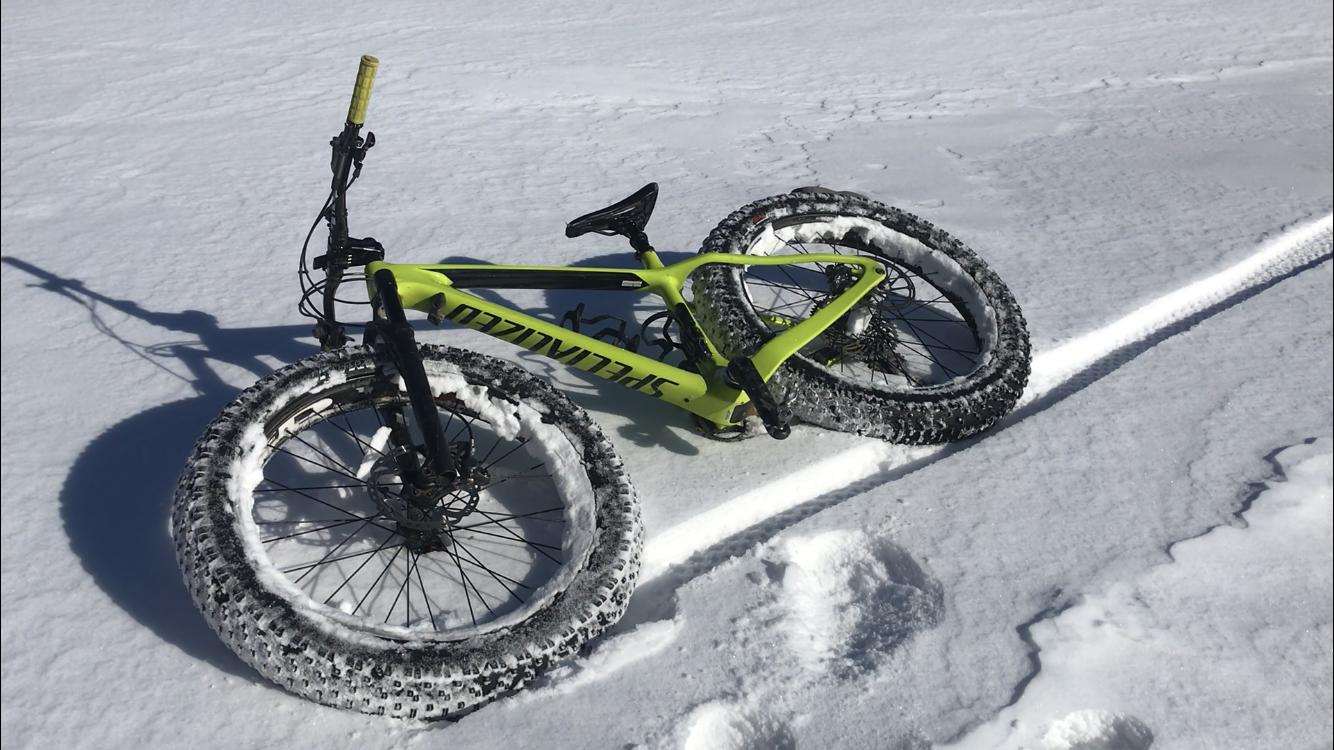 A bright yellow fat bike resting on a snowy surface, with large tires leaving tracks in the snow. The bike is positioned on its side, showcasing its distinctive wide tires designed for winter riding. Mariposa Fat Bike Trails mountain bike trail.
