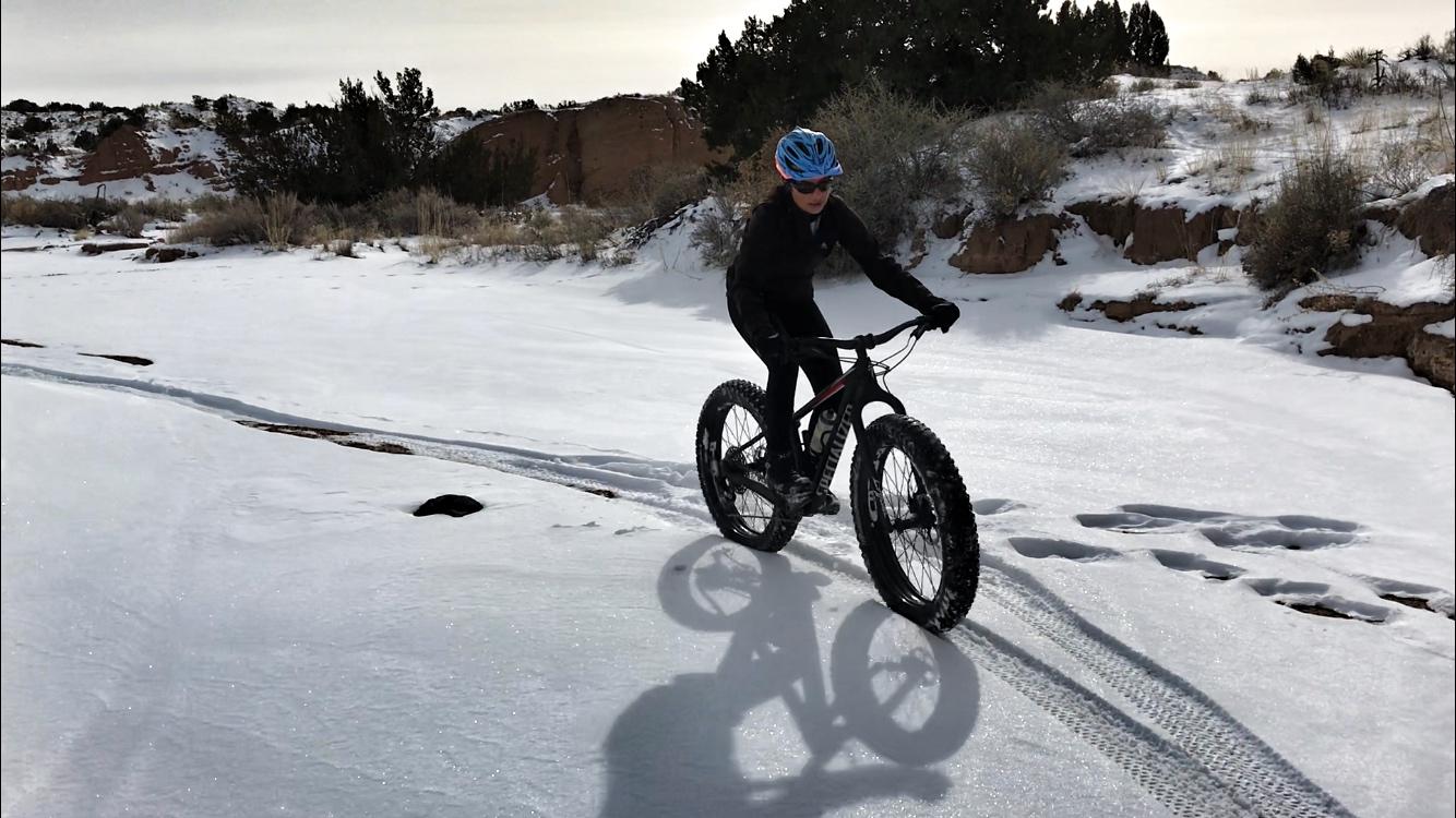 A person riding a fat tire bicycle on snowy ground, leaving tire tracks in the snow. The landscape features sparse vegetation and rocky terrain under a cloudy sky. Mariposa Fat Bike Trails mountain bike trail.