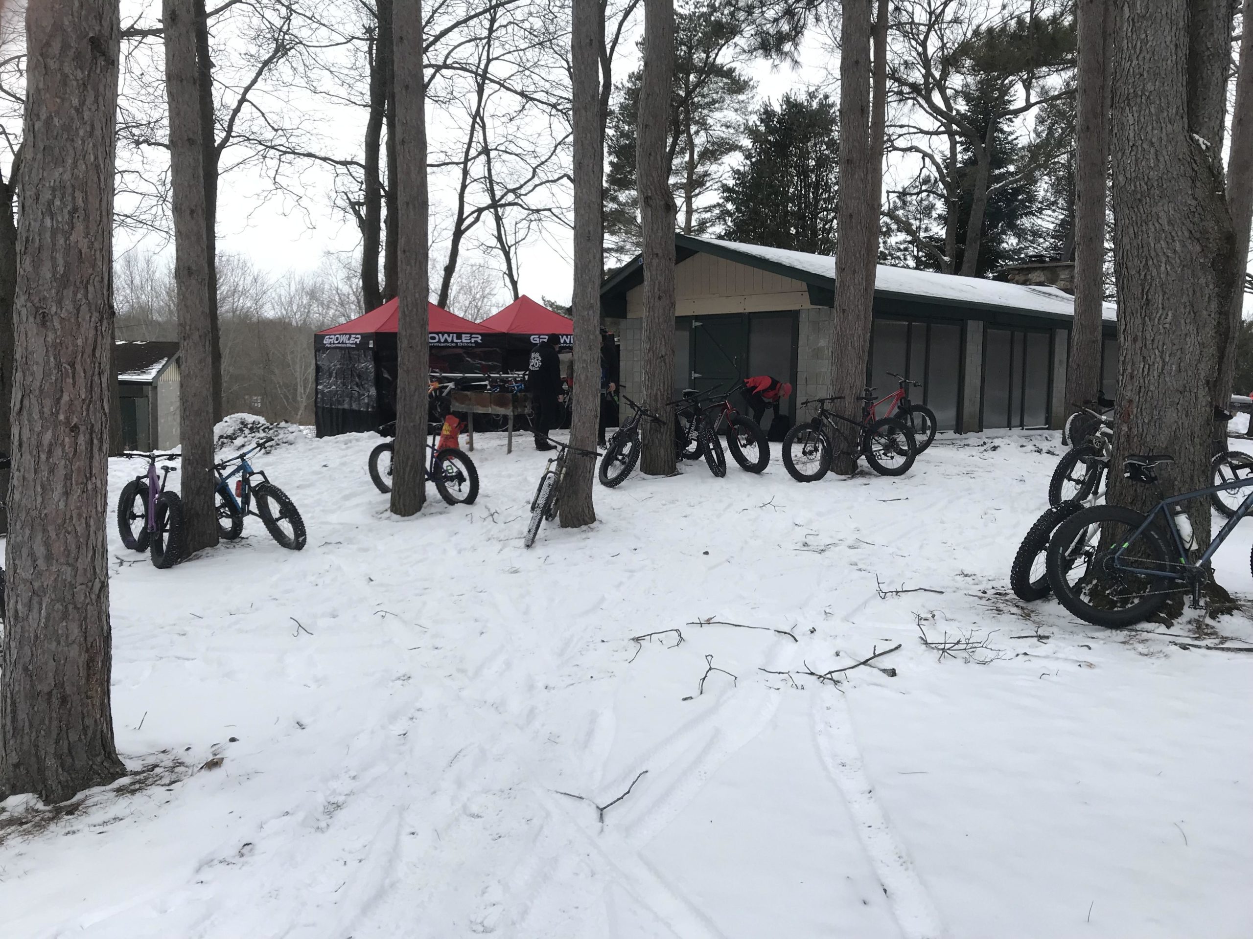 A winter scene featuring several fat bikes parked on snow-covered ground near a cabin. Two red tents are set up nearby, indicating an outdoor biking event, while trees surround the area. Snow blankets the ground, creating a serene and wintry atmosphere. Genesee County Park mountain bike trail.