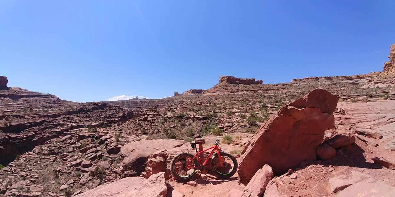 A bright red mountain bike parked on rocky terrain under a clear blue sky, with expansive desert landscapes and distant rock formations in the background. Amasa Back Area mountain bike trail.