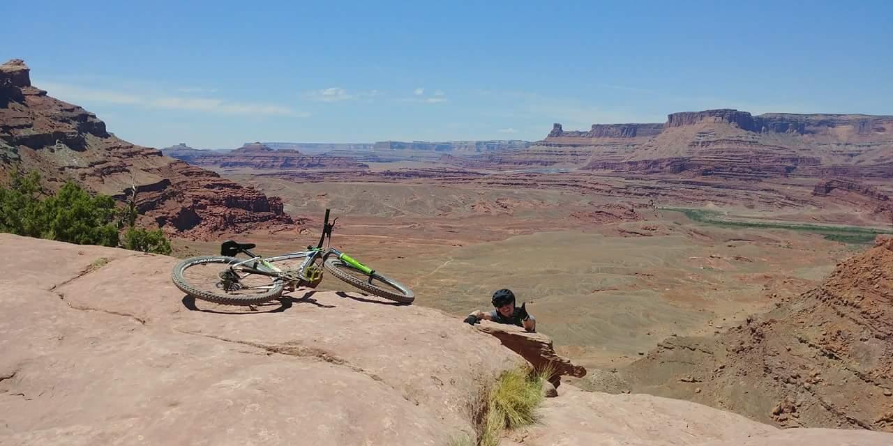 A mountain biker rests near the edge of a rocky cliff, with a bicycle lying on the ground beside him. The background showcases a vast canyon landscape with layered red rock formations under a clear blue sky. Amasa Back Area mountain bike trail.
