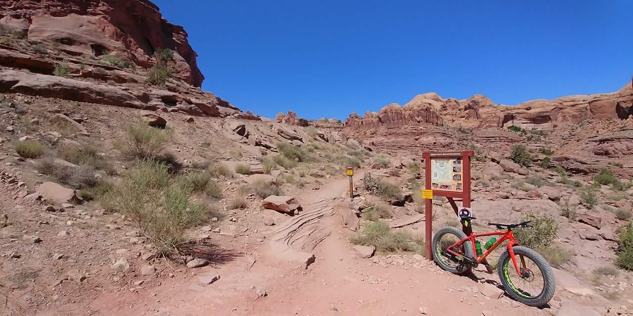 A dirt biking trail surrounded by rocky terrain and red rock formations under a clear blue sky. A brightly colored bicycle is parked next to a trail sign that provides information about the area. Sparse vegetation is scattered along the path, typical of a desert environment. Amasa Back Area mountain bike trail.