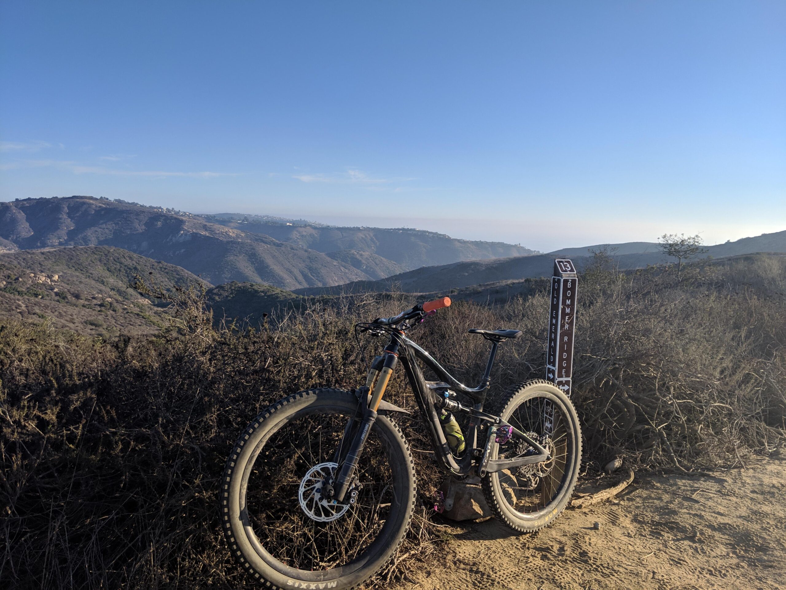 Ibis Mojo HD4: A mountain bike resting on a dirt trail with a scenic view of rolling hills and valleys in the background. A trail sign labeled "Bommer Ridge" is visible alongside the bike, with clear skies above and sparse vegetation in the foreground.