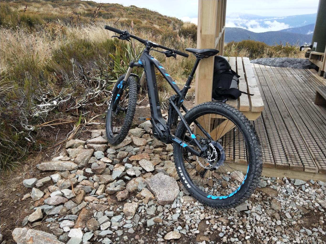 Focus Focus Jam Plus Ltd: A mountain bike is leaning against a rocky area near a wooden bench on a hillside. The background features grassy vegetation and distant mountains under a cloudy sky. A small black backpack is placed on the bench.