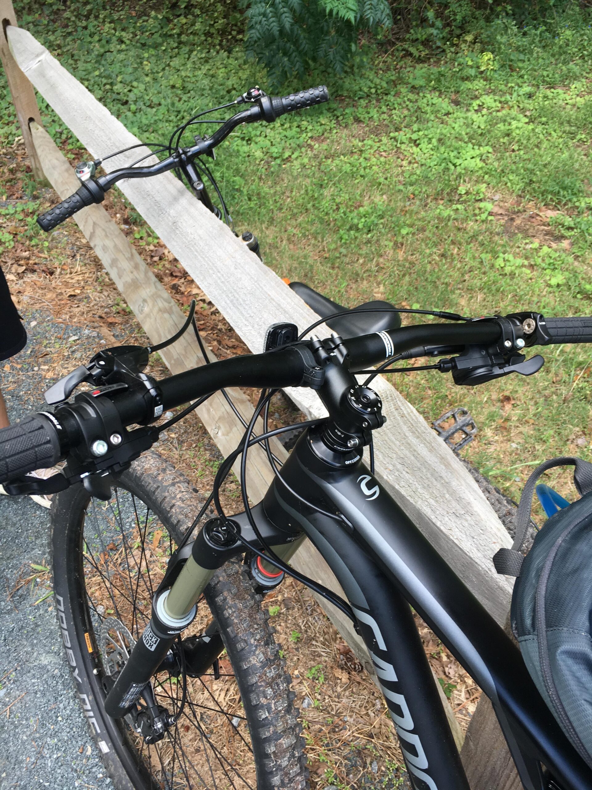 Cannondale Habit: Close-up view of the handlebars and front fork of a black mountain bike, positioned near a wooden fence and surrounded by lush greenery. The image emphasizes the bike's grip and gear shifters, with a focus on the textured handlebar grips and a portion of the bike frame.
