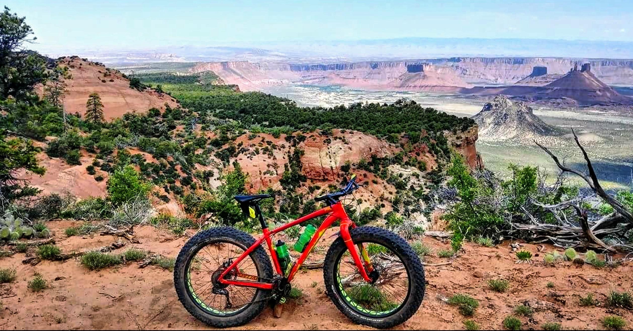 A bright red fat bike resting on a rocky trail overlooking a vast open landscape, featuring red rock formations and lush greenery in the foreground, with distant hills and mesas under a clear blue sky. Porcupine Rim mountain bike trail.
