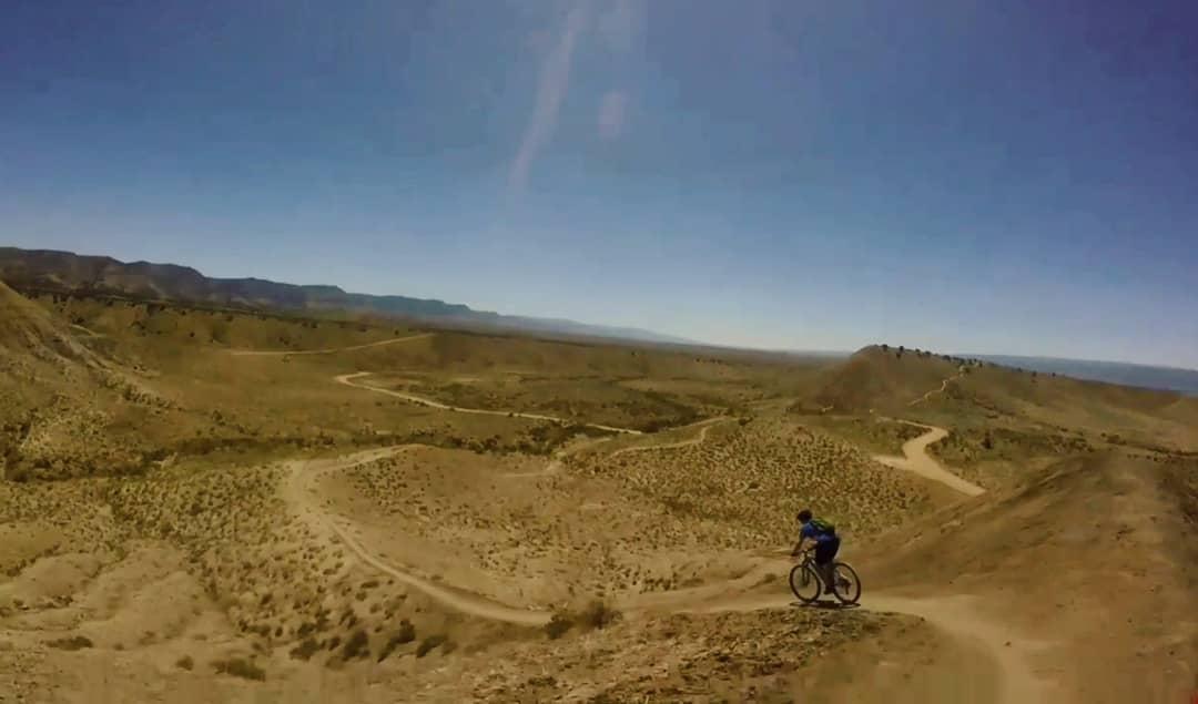 A mountain biker rides along a winding dirt trail in a vast, arid landscape under a clear blue sky. The terrain features rolling hills and sparse vegetation, showcasing a remote, rugged environment ideal for outdoor adventure. 18 Road Trails / North Fruita Desert mountain bike trail.