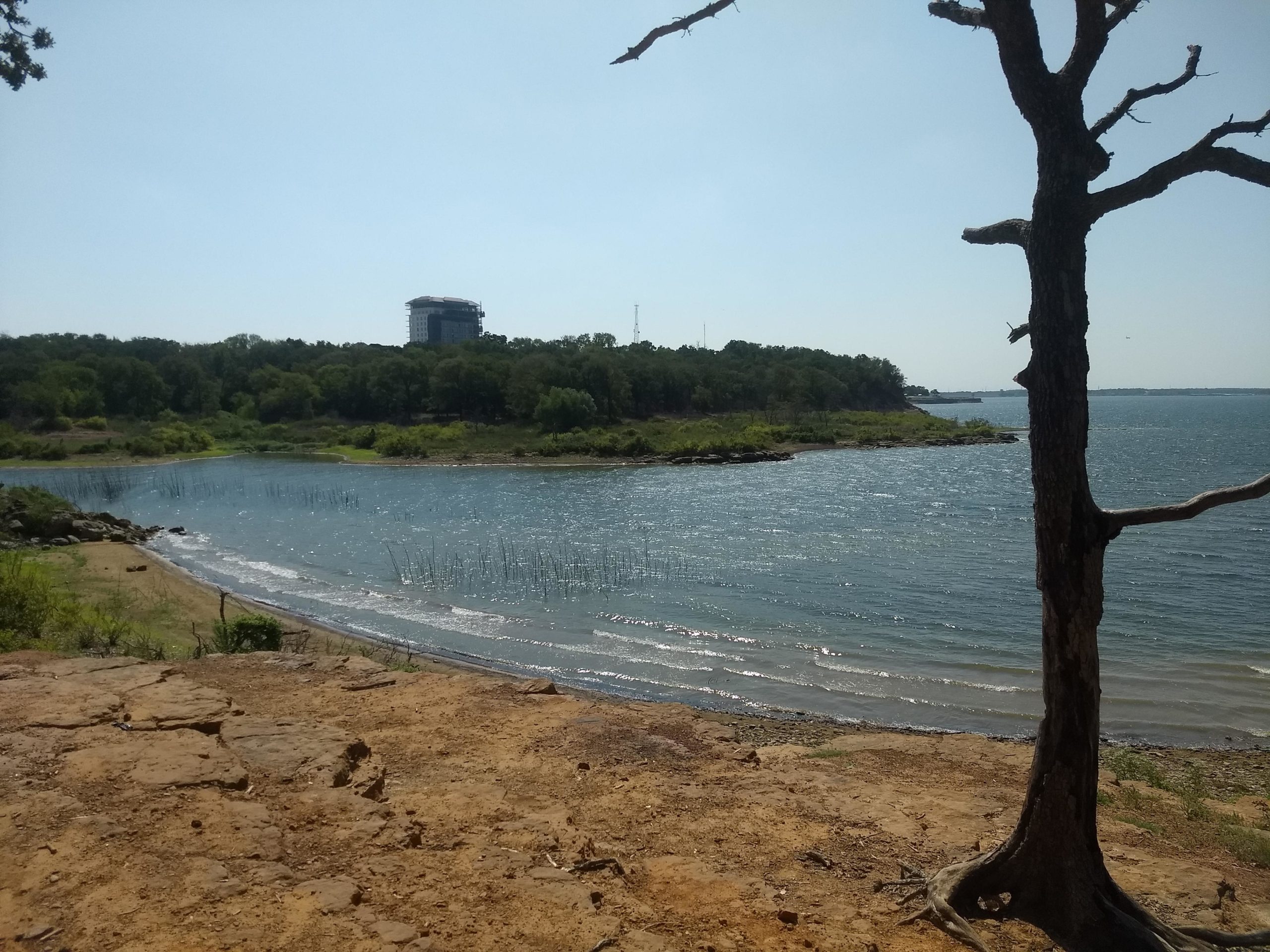 A scenic view of a lake with a sandy shoreline and gentle waves. In the background, a wooded area is visible, along with a partially constructed building situated among the trees. A leafless tree stands in the foreground, adding to the natural setting under a clear blue sky. Northshore Trail mountain bike trail.