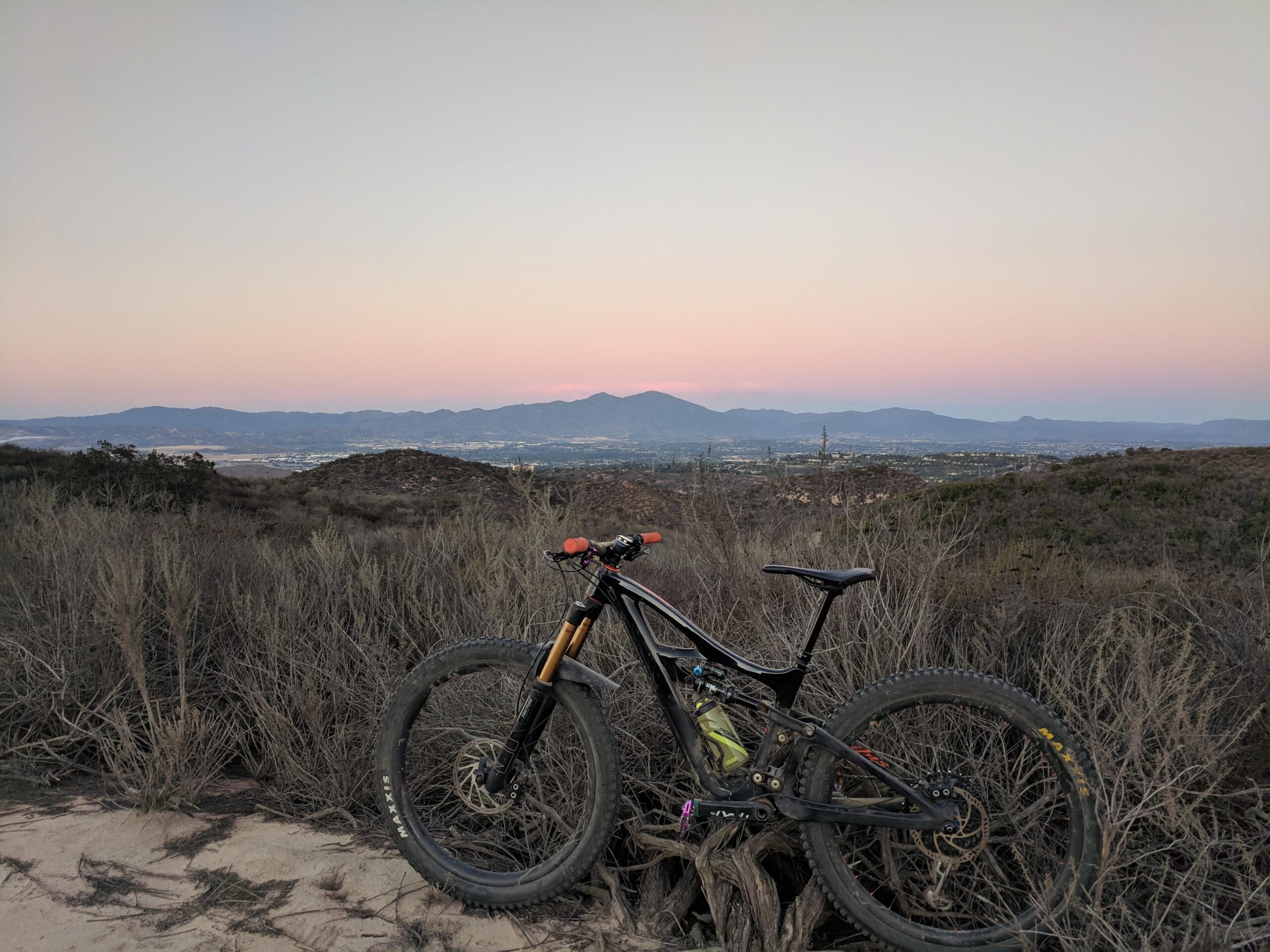 Ibis Mojo HD4: A mountain bike rests against a bush on a hillside, with a panoramic view of a valley and distant mountains under a pastel sunset sky. The foreground shows the bike's front wheel and frame, while the background captures rolling hills and a fading light.