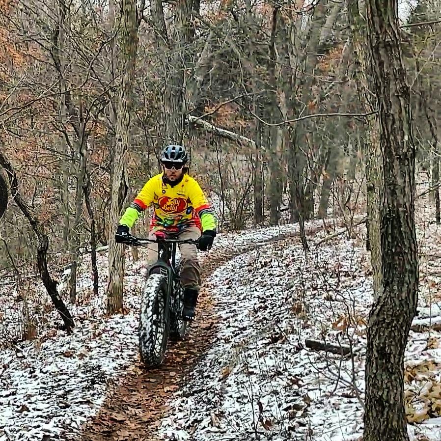 A person riding a fat-tire bicycle on a snowy forest trail, surrounded by bare trees and fallen leaves. The cyclist is wearing a bright yellow and multicolored jersey with gloves and a helmet. DTE Energy Foundation Trail mountain bike trail.