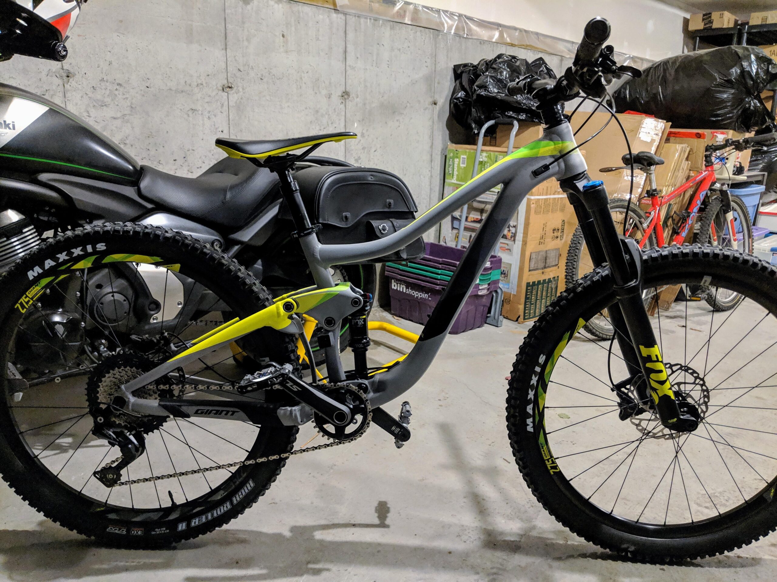 Giant Trance 2: A close-up view of a modern mountain bike positioned in a garage. The bike features a gray and yellow frame, with notable components like a black and yellow fork labeled "FOX" and "MAXXIS" tires. In the background, a motorcycle and various items are visible, contributing to the cluttered garage environment.
