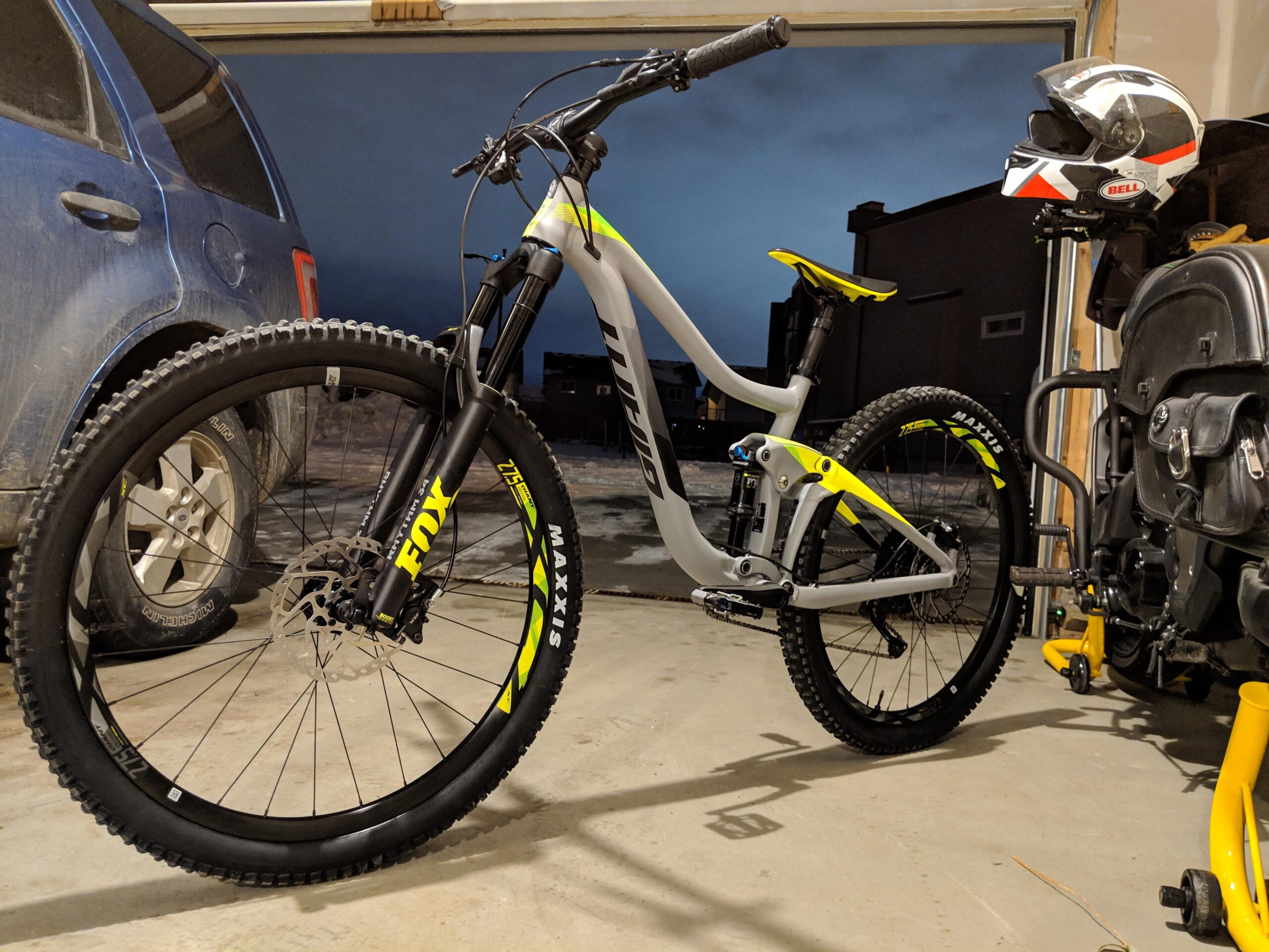 Giant Trance 2: A mountain bike with a gray frame, yellow accents, and black tires is positioned in a garage. A blue SUV is parked in the background, and a helmet is mounted on a wall hook nearby. The lighting suggests early evening, with a dark blue sky visible outside.