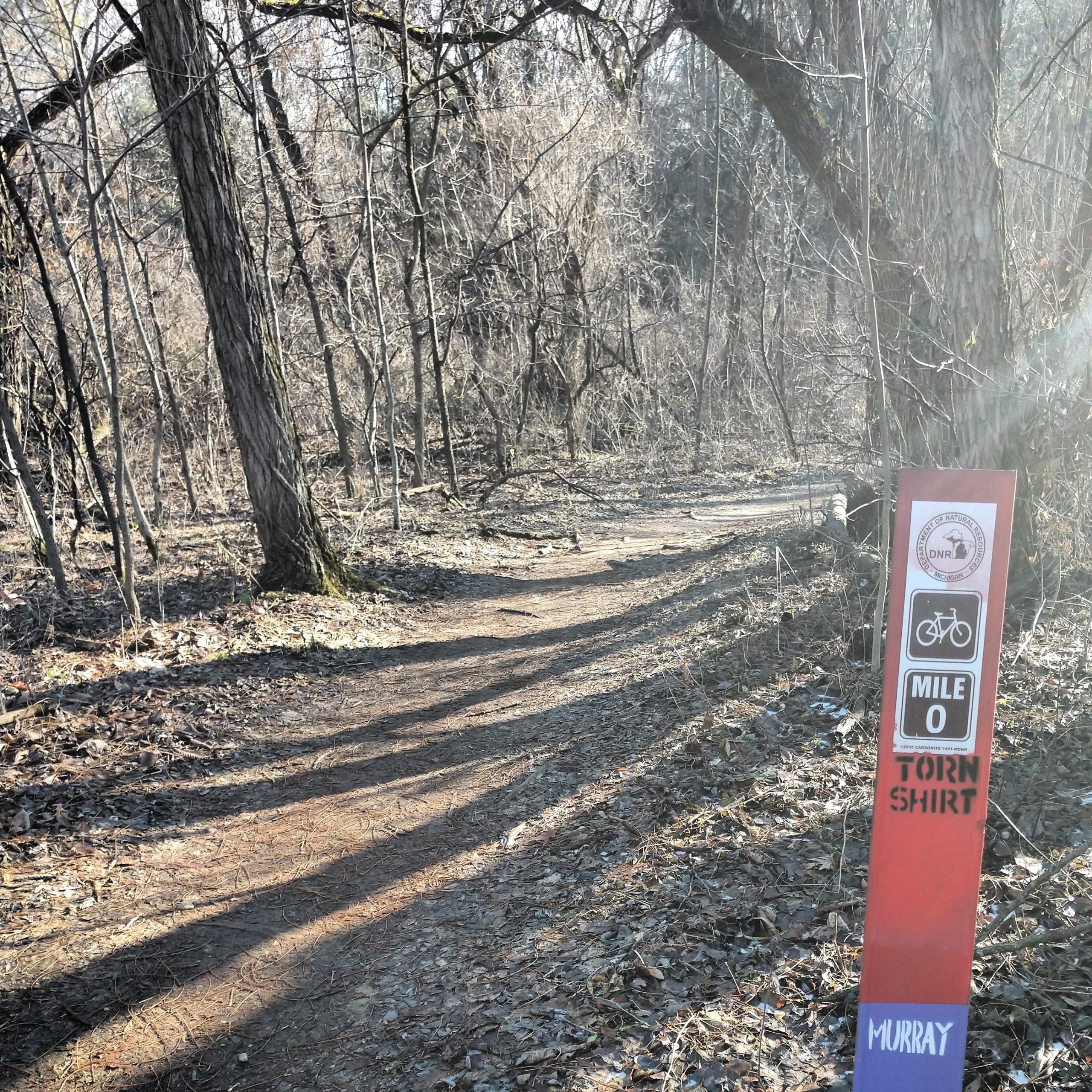 A dirt path winding through a wooded area, with bare trees in the background. On the right side, there's a trail marker indicating "MILE 0" and "TORN SHIRT," with symbols for biking. Sunlight casts long shadows on the ground, creating a serene outdoor atmosphere. Brighton Rec Area mountain bike trail.