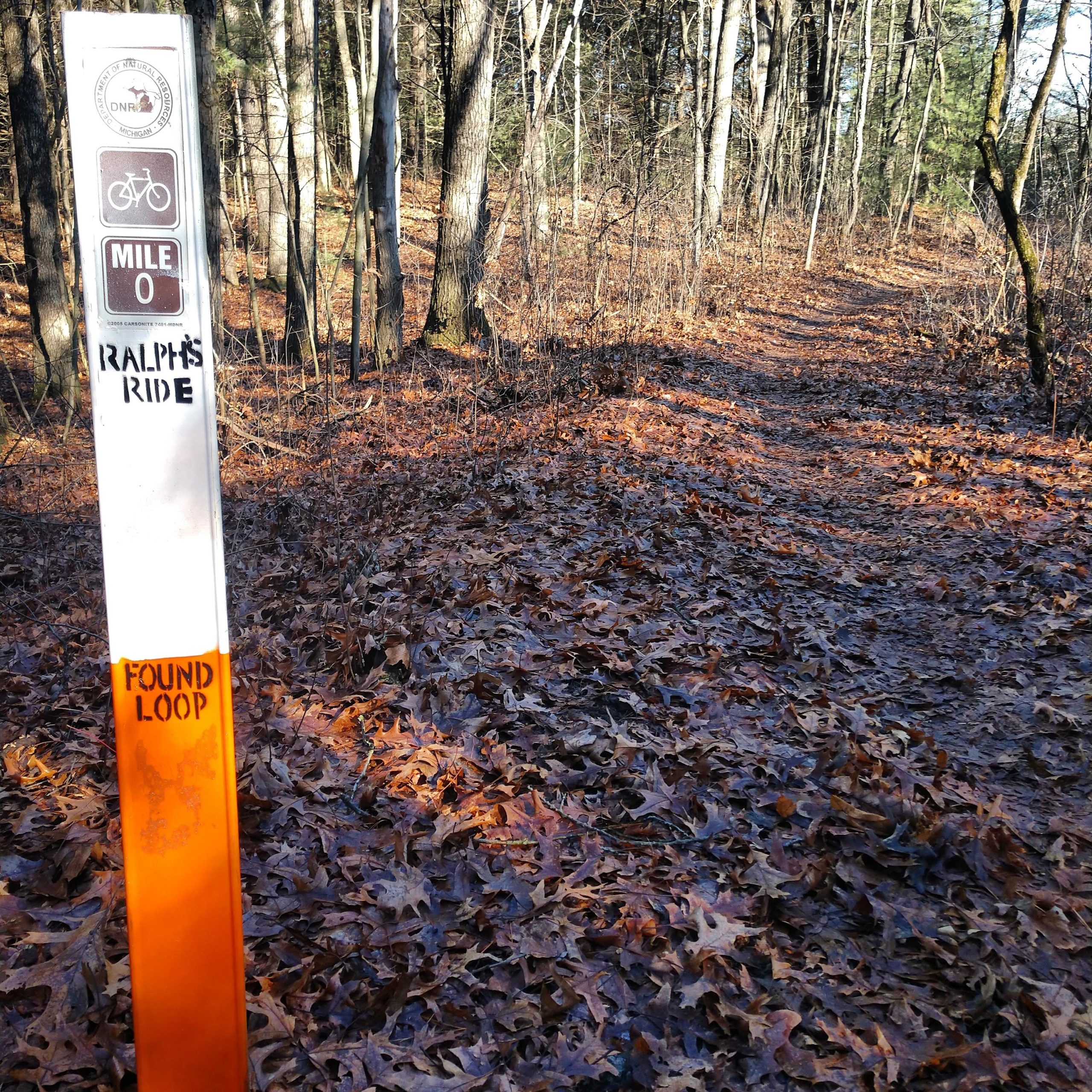 A trail marker for a bike path indicates "Mile 0," labeled "Ralph's Ride," with a section labeled "Found Loop." The surroundings are forested with bare trees and a path covered in fallen leaves. Brighton Rec Area mountain bike trail.