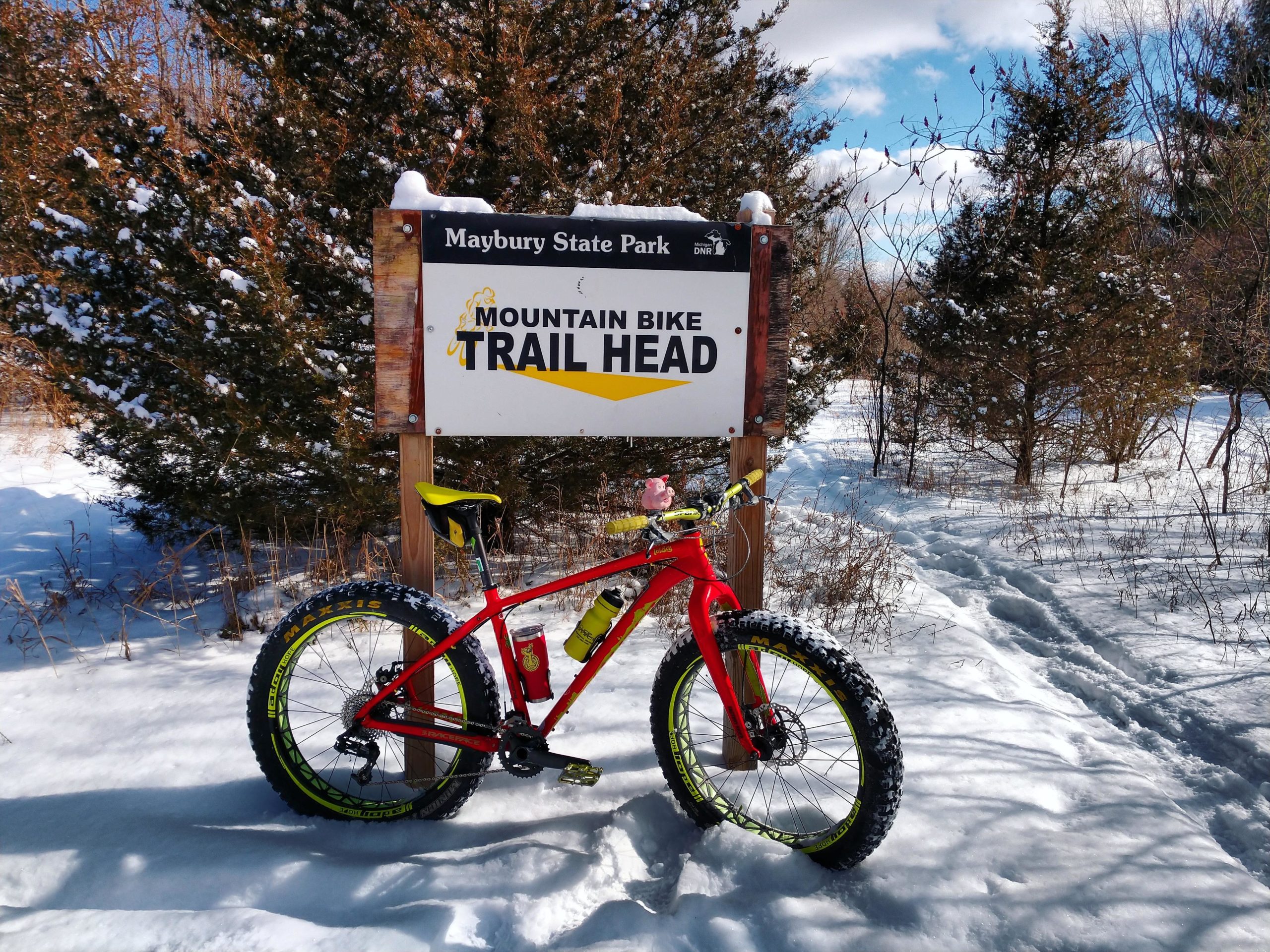 A red fat bike parked next to a trailhead sign labeled "Mountain Bike Trail Head" at Maybury State Park, with a snowy landscape and trees in the background. Maybury mountain bike trail.