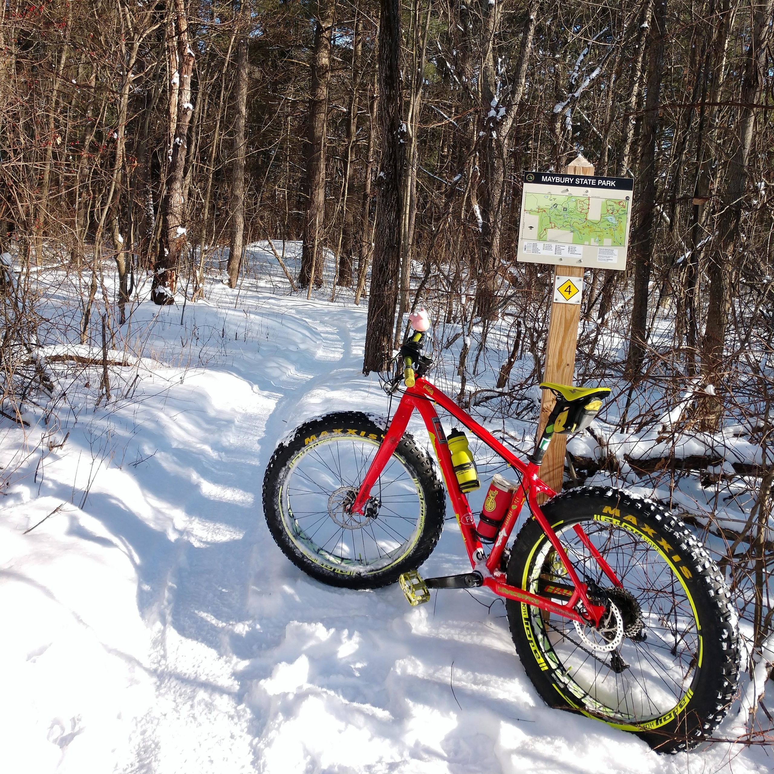 A vibrant red fat bike parked on a snow-covered trail in Maybury State Park, alongside a map sign displaying park information. Surrounding the bike are trees with snow-dusted branches, creating a serene winter atmosphere. Maybury mountain bike trail.