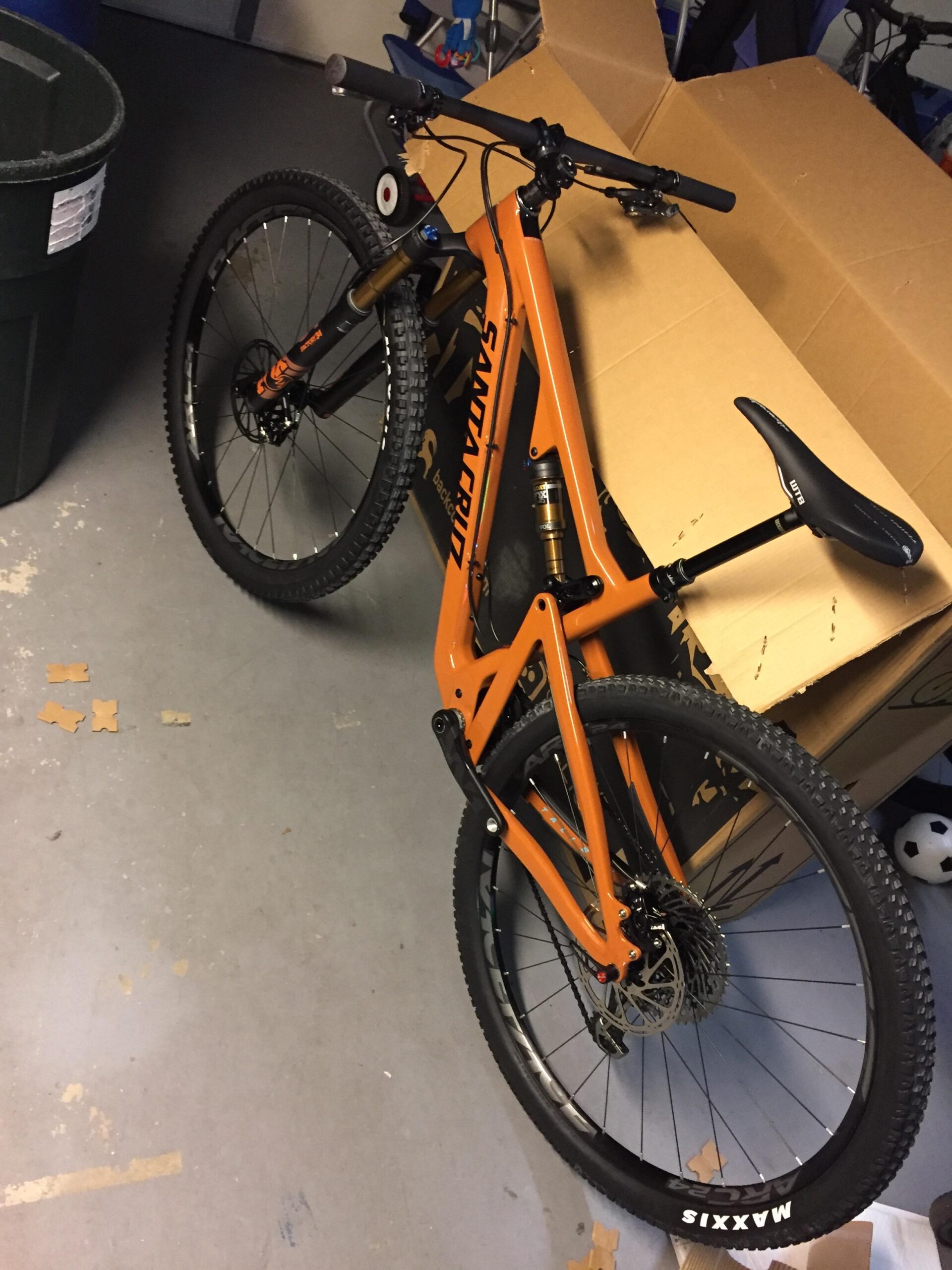 Santa Cruz Santa Cruz Tallboy CC: A close-up view of an orange mountain bike leaning against a cardboard box in a garage. The bike features thick tires labeled 'Maxxis' and a suspension system. A trash can is visible in the background, along with a soccer ball and some scattered cardboard pieces on the floor.