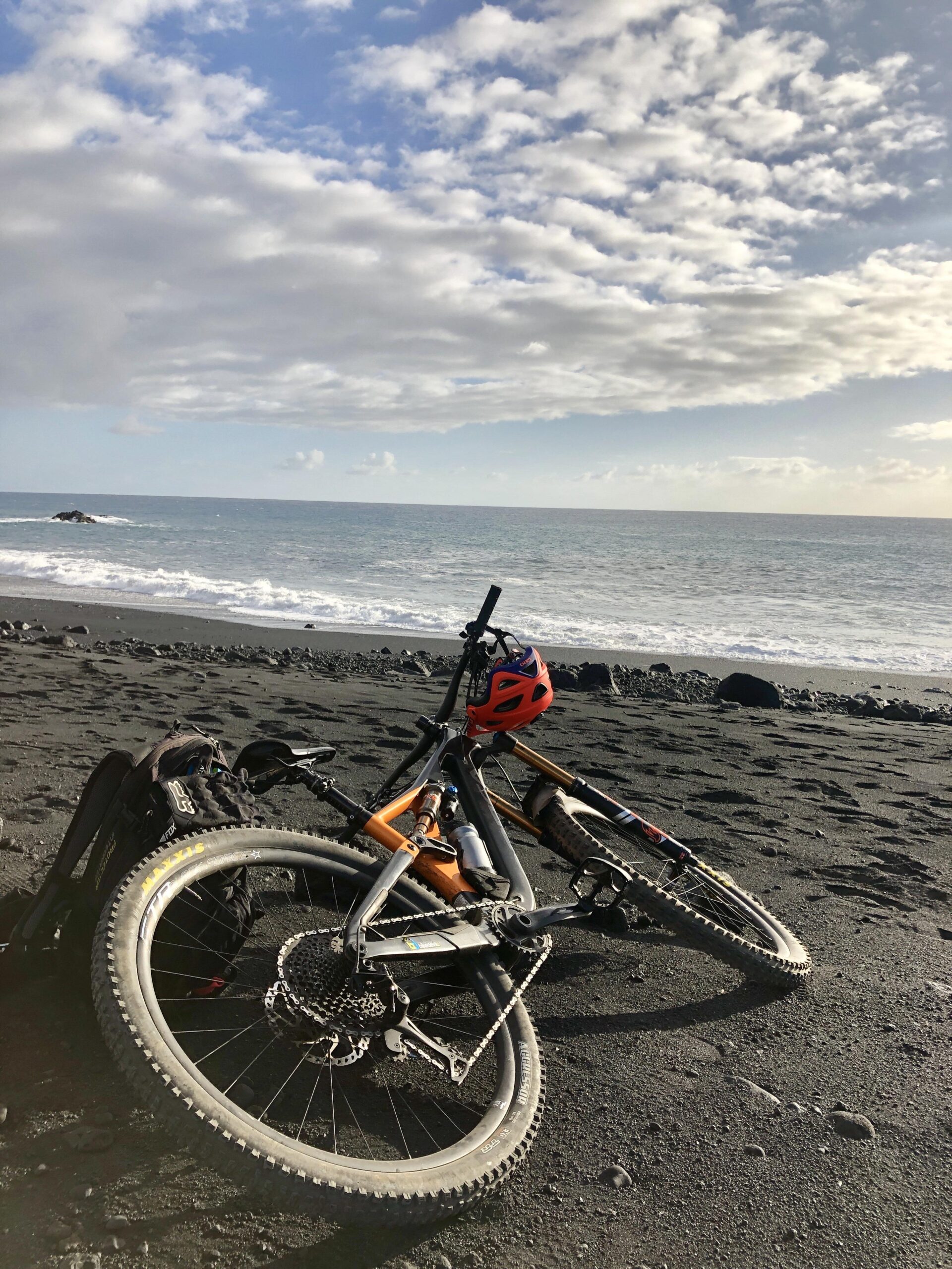 Orbea Rallon: Two mountain bikes resting on a black sand beach with gentle waves in the background. One bike is orange and gray, and the other is gray with a visible water bottle. A bright red helmet sits on one of the bikes. The sky is partly cloudy, creating a serene atmosphere at the seaside.