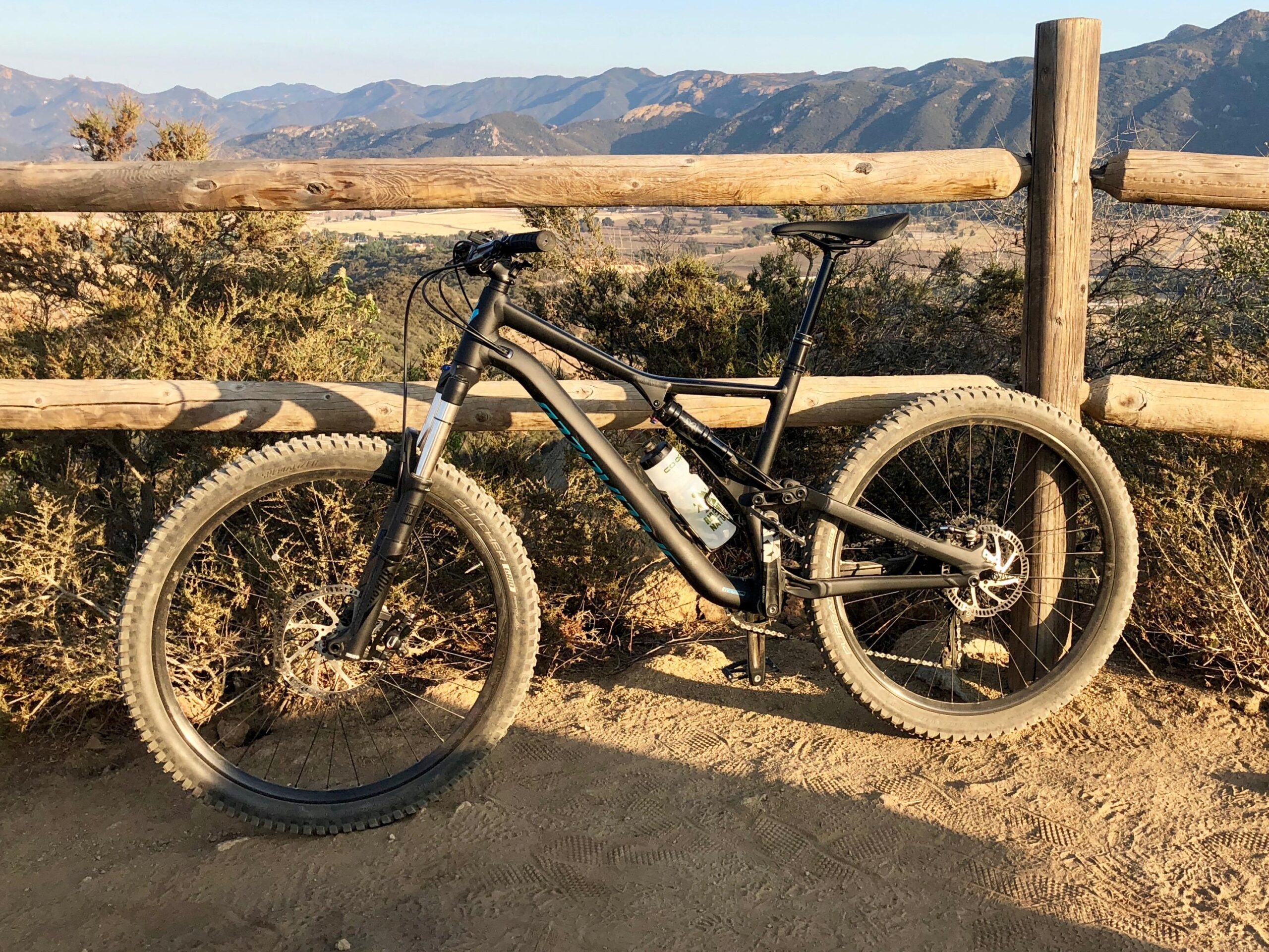 Specialized Stumpjumper: A black mountain bike resting against a wooden fence on a dirt trail, with a backdrop of mountains and greenery under clear skies.