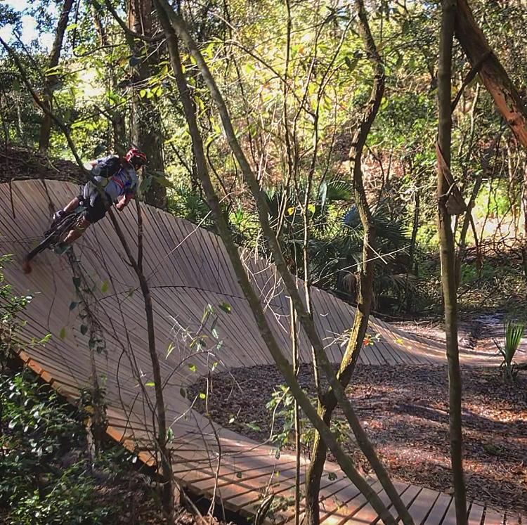A mountain biker navigating a wooden pump track through a dense forest, surrounded by trees and greenery. The cyclist is leaning into a curve of the track, showcasing an action shot in a natural setting. Alafia River State Park mountain bike trail.