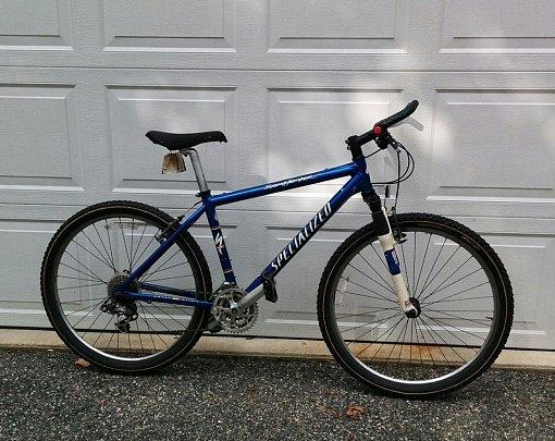 Specialized Stumpjumper: A blue Specialized mountain bike is leaning against a white garage door. The bike features thick tires, a suspension fork, and a straightforward gear setup, suitable for off-road riding. The scene is outdoors, with a gravel surface beneath the bike.